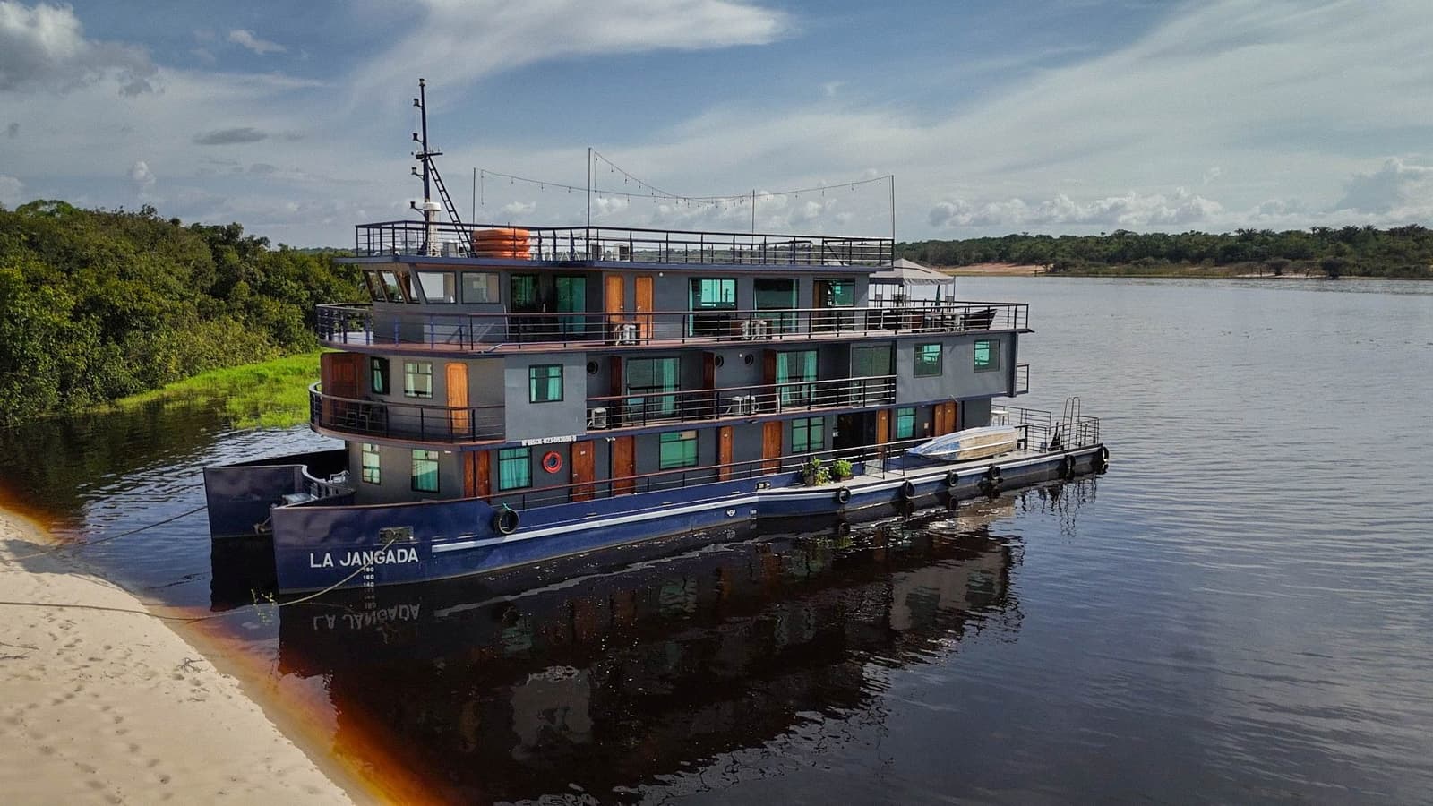Tre ponti, dodici cabine e un fiume quasi nero: il viaggio comincia qui. L'imbarcazione La Jangada ormeggiata sulle acque scure del Rio Negro, con lo scafo in acciaio a tre ponti e le cabine in legno riflesse nel fiume e la fitta foresta amazzonica sulla riva opposta