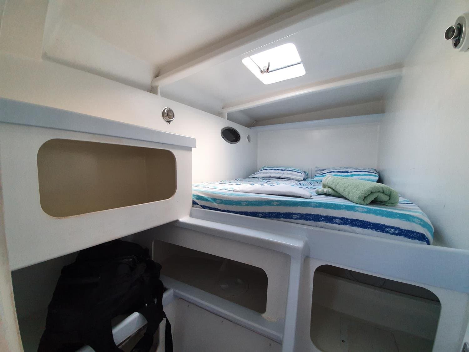 Small but yours, a cozy cabin with a skylight beats a hotel room any day. Compact double berth inside a liveaboard Abrolhos catamaran cabin, made up with blue-striped bedding, a folded green towel, a small oval porthole, and a roof hatch letting in natural light