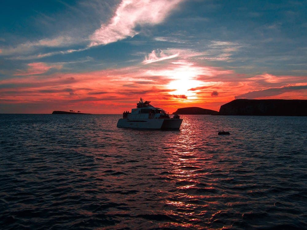 Sundowner at sea, no Wi-Fi, no complaints. A white catamaran silhouetted against a vivid red and orange sunset over the Abrolhos archipelago, with two low volcanic islands visible on the horizon and the sun's reflection stretching across calm open water — the liveaboard Abrolhos experience at the end of the day.
