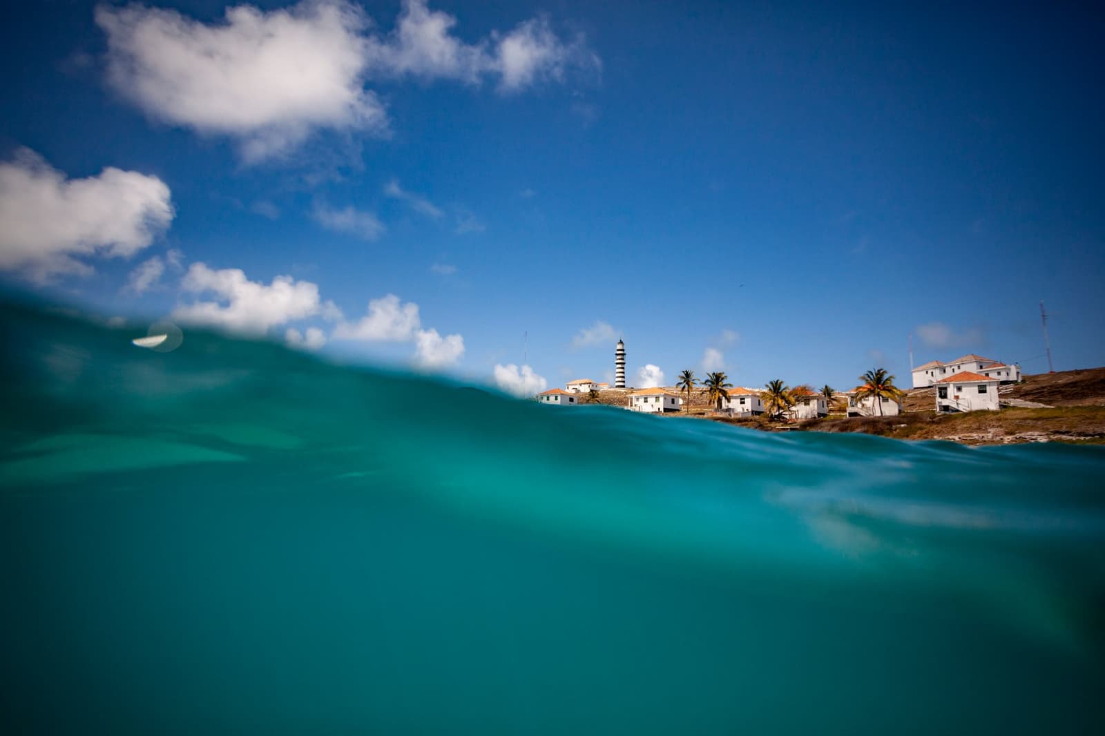 Half ocean, half island. The lighthouse has been keeping watch long before we thought to swim past it. Photo: FOTO_GabiCarrera Split-level shot of the liveaboard Abrolhos experience: turquoise water fills the foreground while the black-and-white striped Abrolhos Lighthouse, white buildings, and palm trees on Santa Barbara Island rise above the waterline under a blue sky.