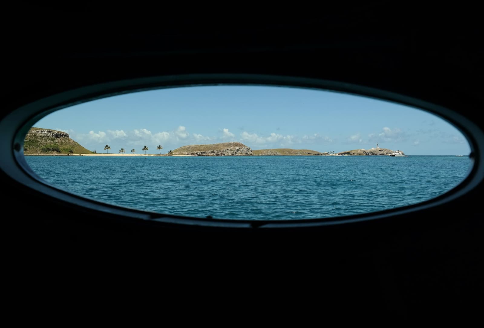 The archipelago, framed and waiting. Your next dive is out there. Photo: Leoaraujo7 View through an oval boat porthole of the liveaboard Abrolhos catamaran, framing rocky volcanic islands with palm trees, a sandy beach, a lighthouse, and turquoise water under a blue sky.