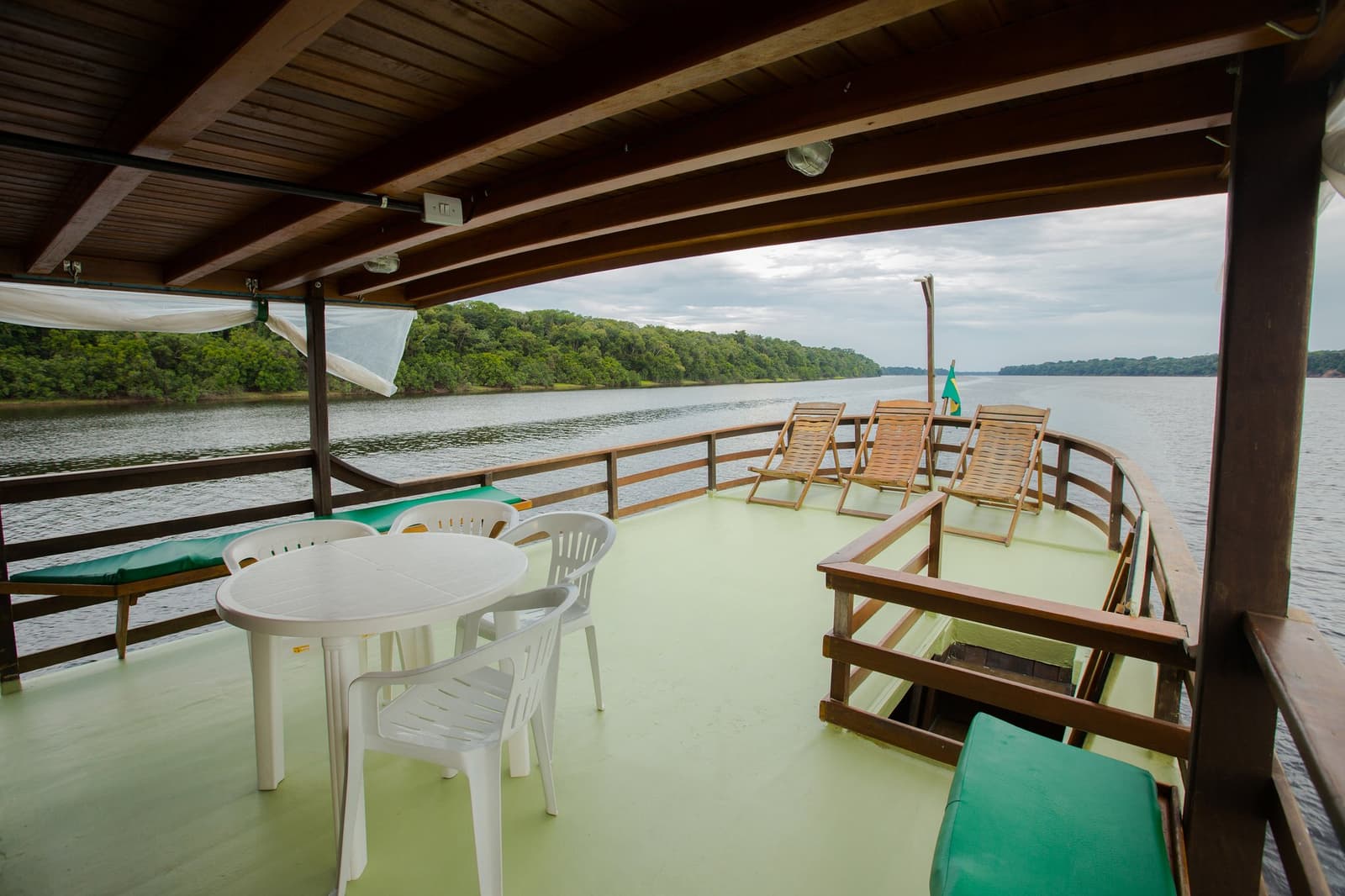 Espreguiçadeira ou mesinha na sombra, o rio cuida do resto. Photo: sitah Convés superior coberto de um barco de cruzeiro pelo rio Amazonas no Brasil, com mesa e cadeiras de plástico branco, espreguiçadeiras de madeira, uma bandeira brasileira e um rio de águas escuras ladeado por densa cobertura de floresta.