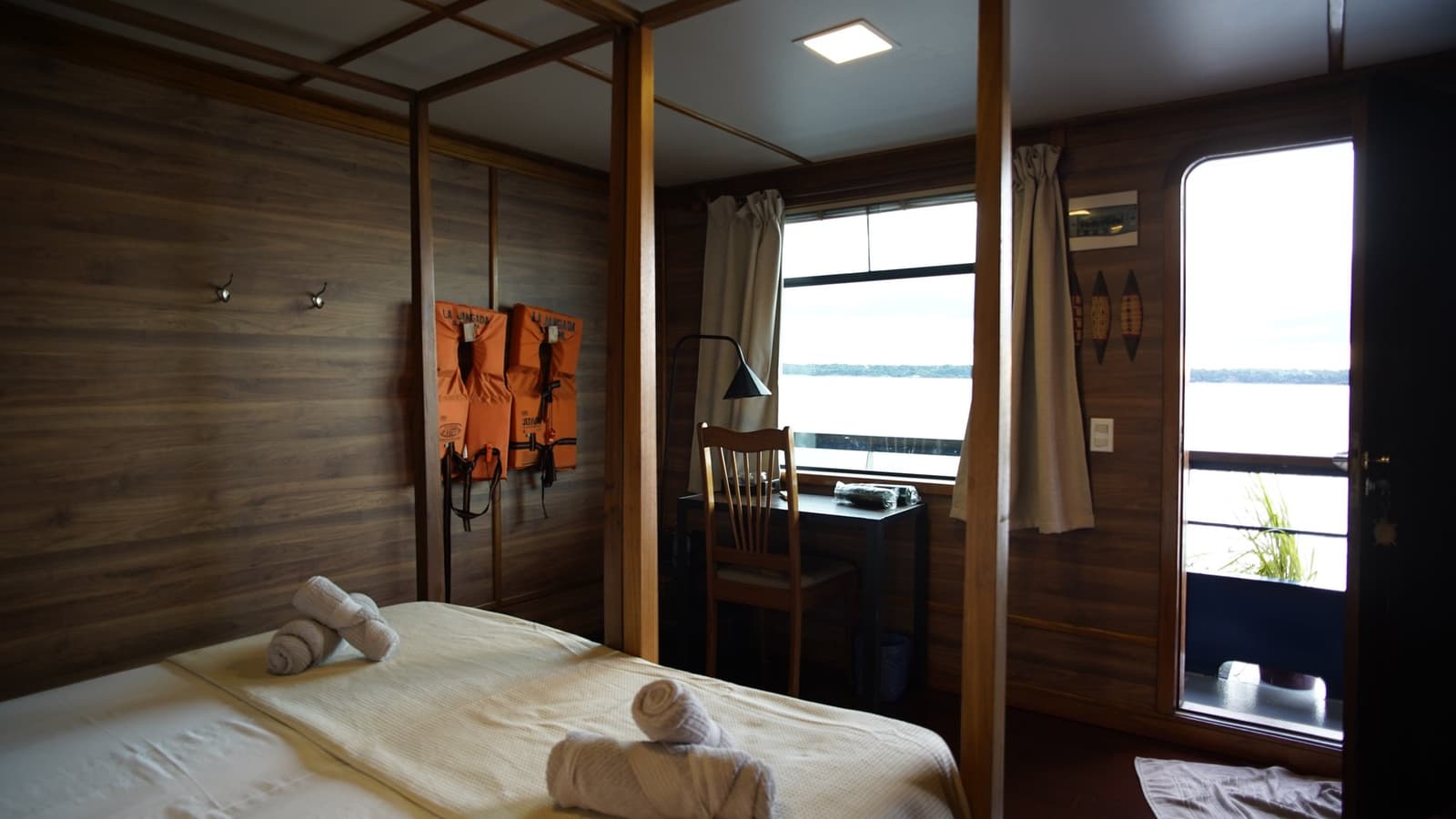 Your room comes with a river on one side and life jackets on the other. Priorities sorted. Wood-panelled cabin on the La Jangada amazon cruise with a double bed dressed in white linens, rolled towels, orange life jackets on the wall, a small desk, and a river-view window.