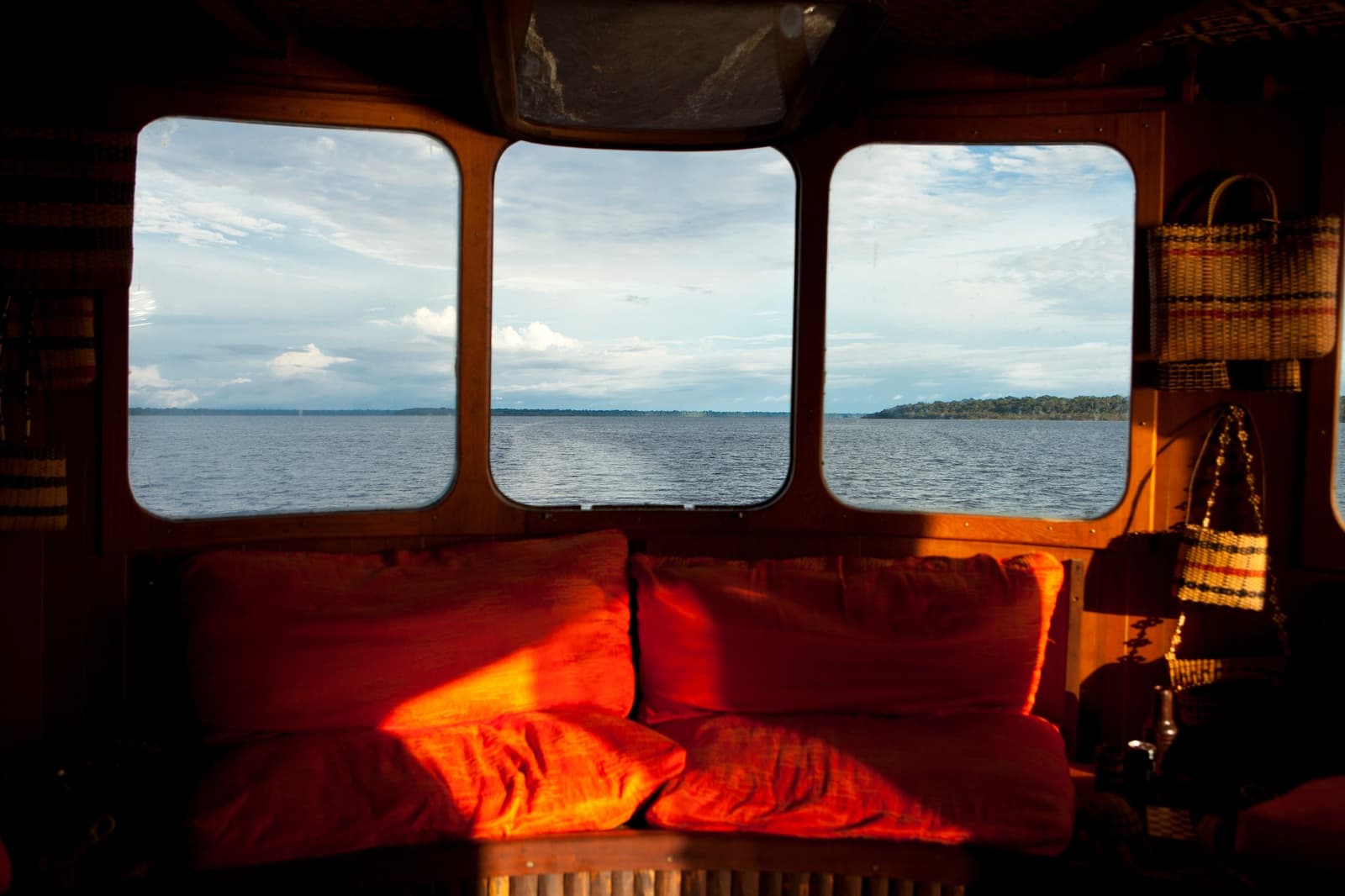 O melhor lugar do barco, sem precisar chegar cedo. Interior de madeira aconchegante de um barco de cruzeiro no Amazonas no Brasil, com banco estofado em almofadas vermelhas e três janelas panorâmicas emoldurando um rio largo sob céu parcialmente nublado, cestas trançadas penduradas à direita