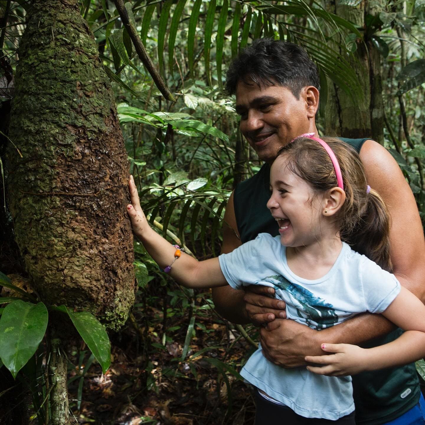 L'albero ha secoli di vantaggio, ma la sua curiosità non conosce limiti. Una bambina sorridente tocca la corteccia di un grande albero della foresta pluviale durante un'escursione guidata della la jangada amazon cruise, tenuta da un adulto sorridente alle sue spalle
