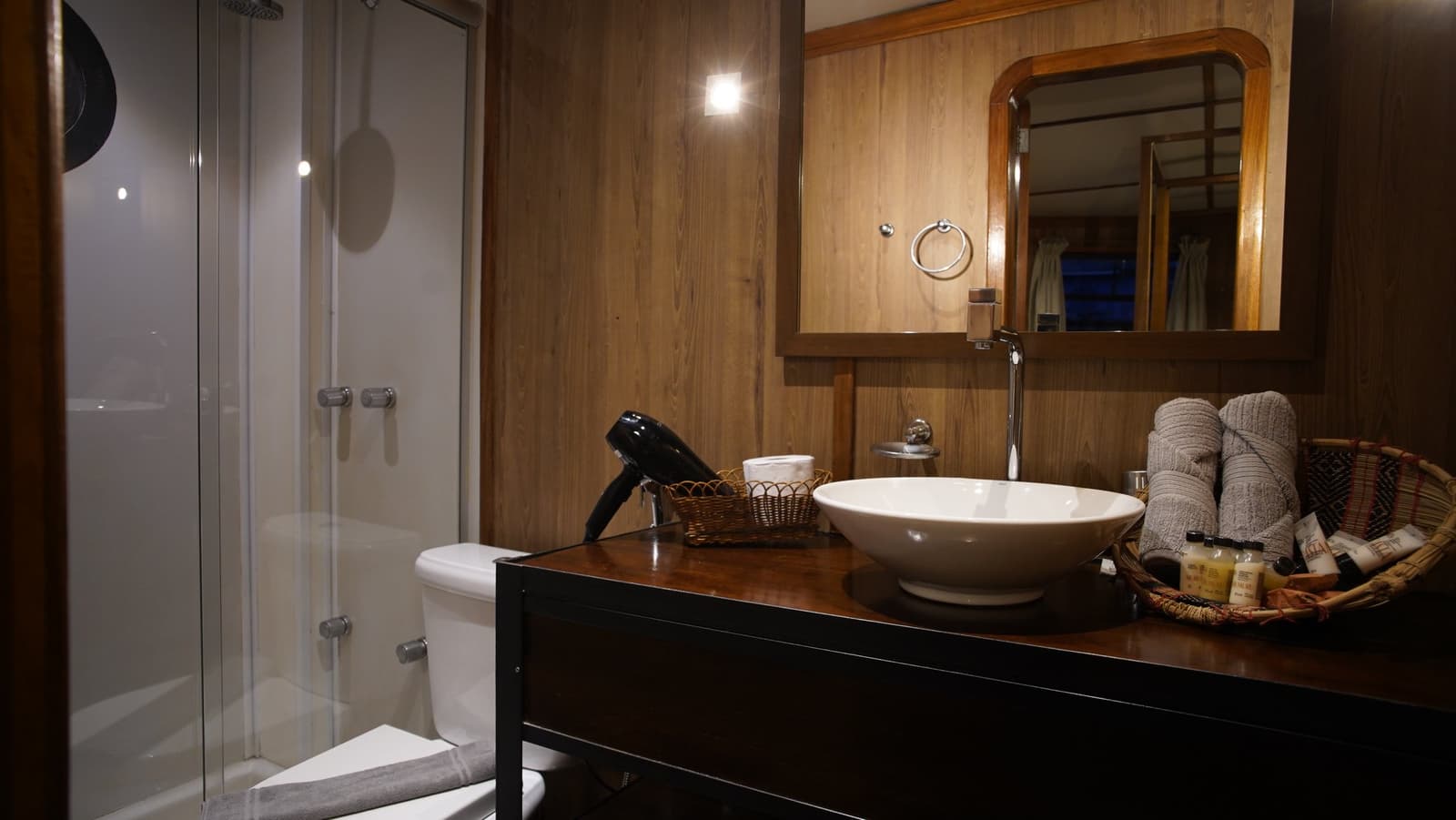 Compact, comfortable, and surprisingly civilised for a boat deep in the jungle. Private en-suite bathroom on the La Jangada Amazon cruise, featuring a vessel bowl sink on a dark wood vanity, rolled grey towels in a wicker basket, a glass-enclosed shower, and warm wood-panelled walls.