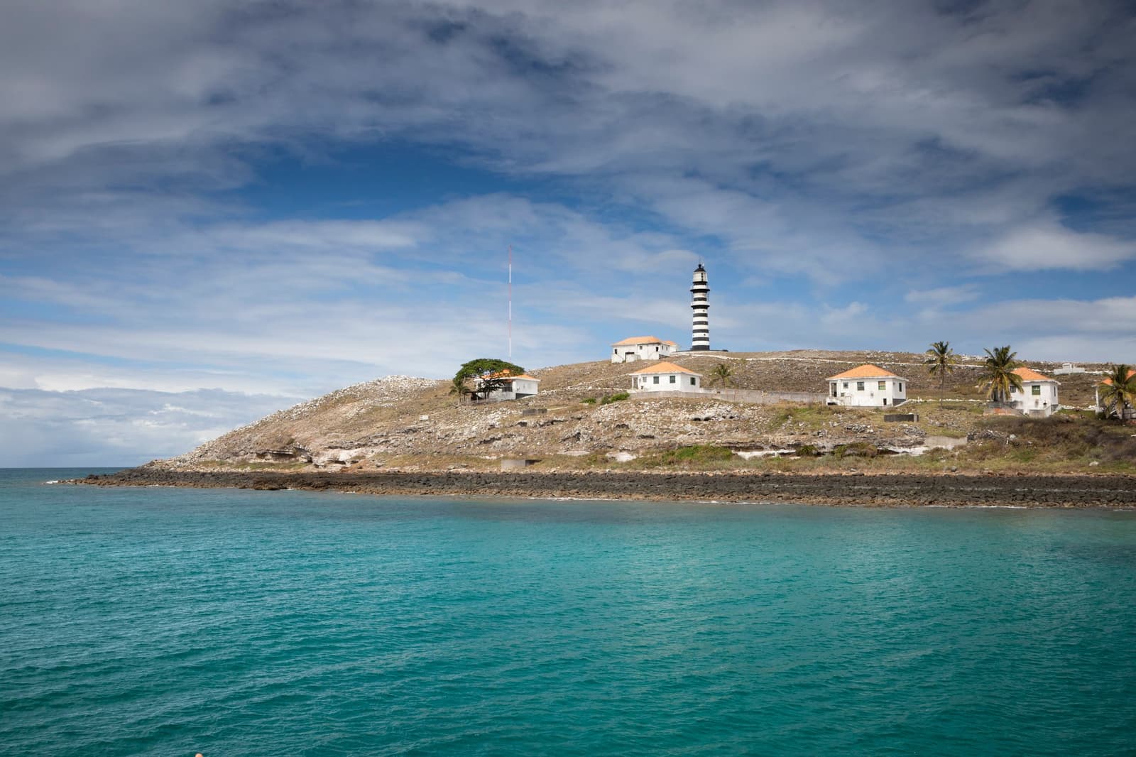 The lighthouse that seabirds use as a landmark and divers as a finishing touch. Photo: FOTO_GabiCarrera A black-and-white striped lighthouse on a rocky hillside above turquoise water, surrounded by low white buildings and scattered palms on the liveaboard Abrolhos route to Santa Barbara Island