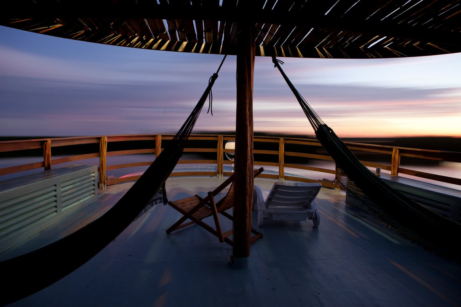 O barco segue, a rede balança, e o crepúsculo toma conta do resto. Duas redes penduradas sob uma cobertura de madeira entrelaçada no deck superior aberto de um barco de cruzeiro pelo rio Amazonas no Brasil, com espreguiçadeiras e uma cadeira dobrável de madeira emoldurando o crepúsculo sobre a planície fluvial