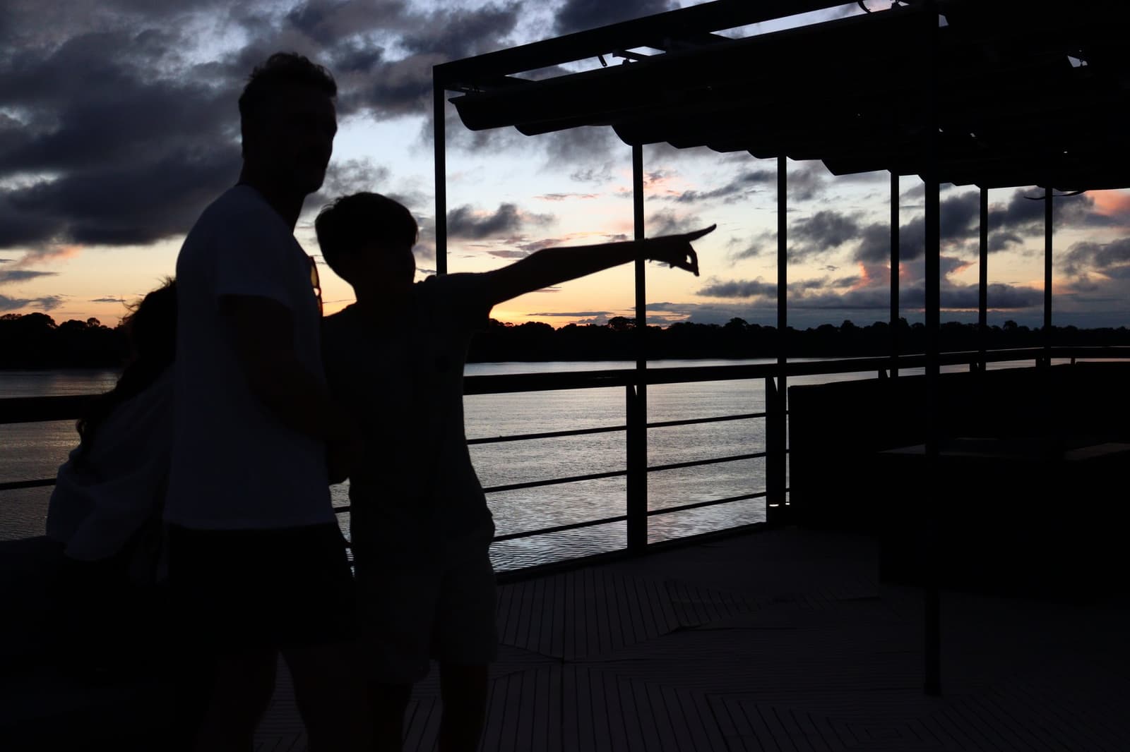 C'è sempre qualcuno che vede qualcosa per primo. È a questo che serve il ponte. Due ospiti in silhouette sul ponte della La Jangada, uno che indica il cielo arancione e viola del tramonto sopra un ampio fiume con rive boscose all'orizzonte