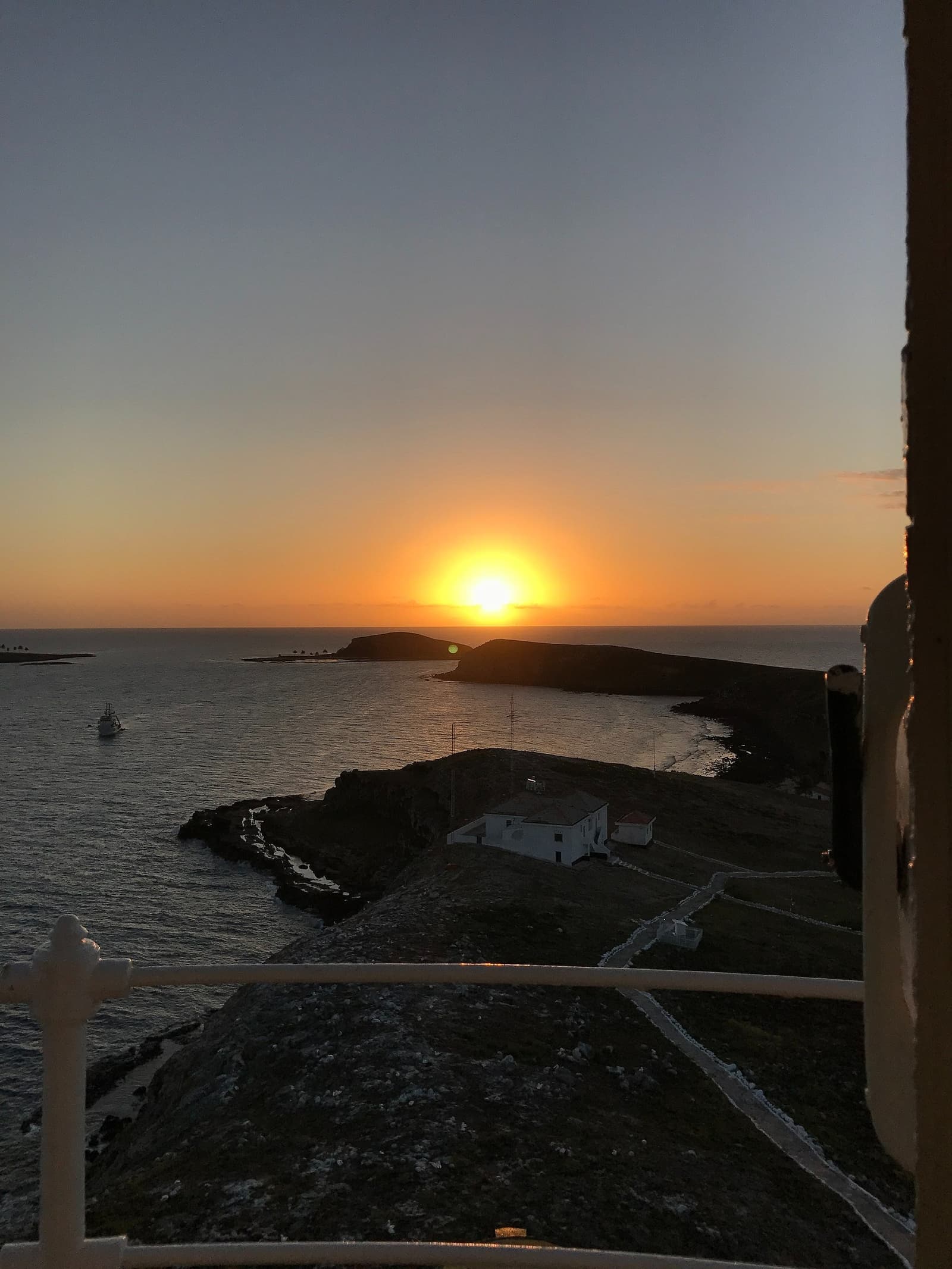 Last light from the top of the lighthouse, the ocean doing the rest. Sunset view from a lighthouse balcony on the liveaboard Abrolhos route, with the sun dipping behind a small volcanic island over a calm bay, a white building below and two vessels on the water