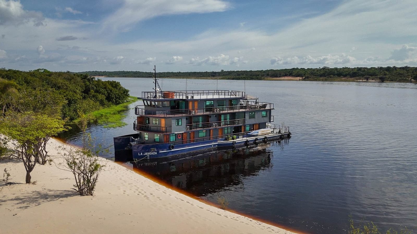 Home for five days, parked somewhere most maps don't bother naming. The La Jangada amazon cruise moored alongside a white sand beach on the dark waters of the Rio Negro, with dense rainforest lining the far bank under a partly cloudy sky