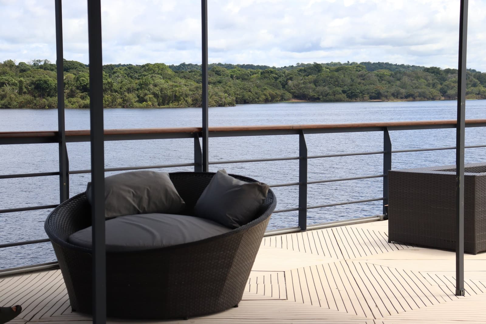 The best office chair you will ever sit in, no question. A cushioned round wicker lounger on the open deck of the La Jangada amazon cruise, with a wide river and dense rainforest canopy visible beyond the railing