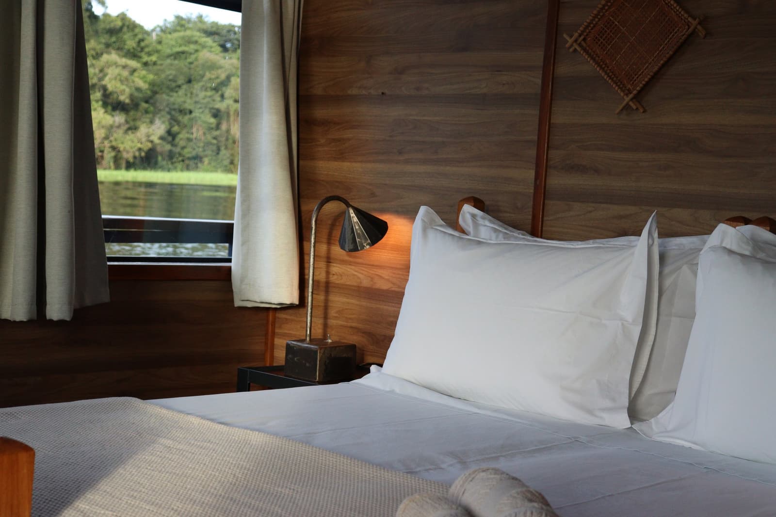 Sleep to the sound of the Rio Negro, wake up to a wall of green. A neatly made double bed with crisp white pillows on the La Jangada amazon river cruise, with a warm wooden headboard, a brass reading lamp, and a window overlooking a green riverbank