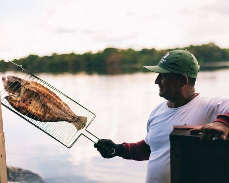 A cook grilling a whole Amazonian fish in a grill basket on the riverbank at sunset