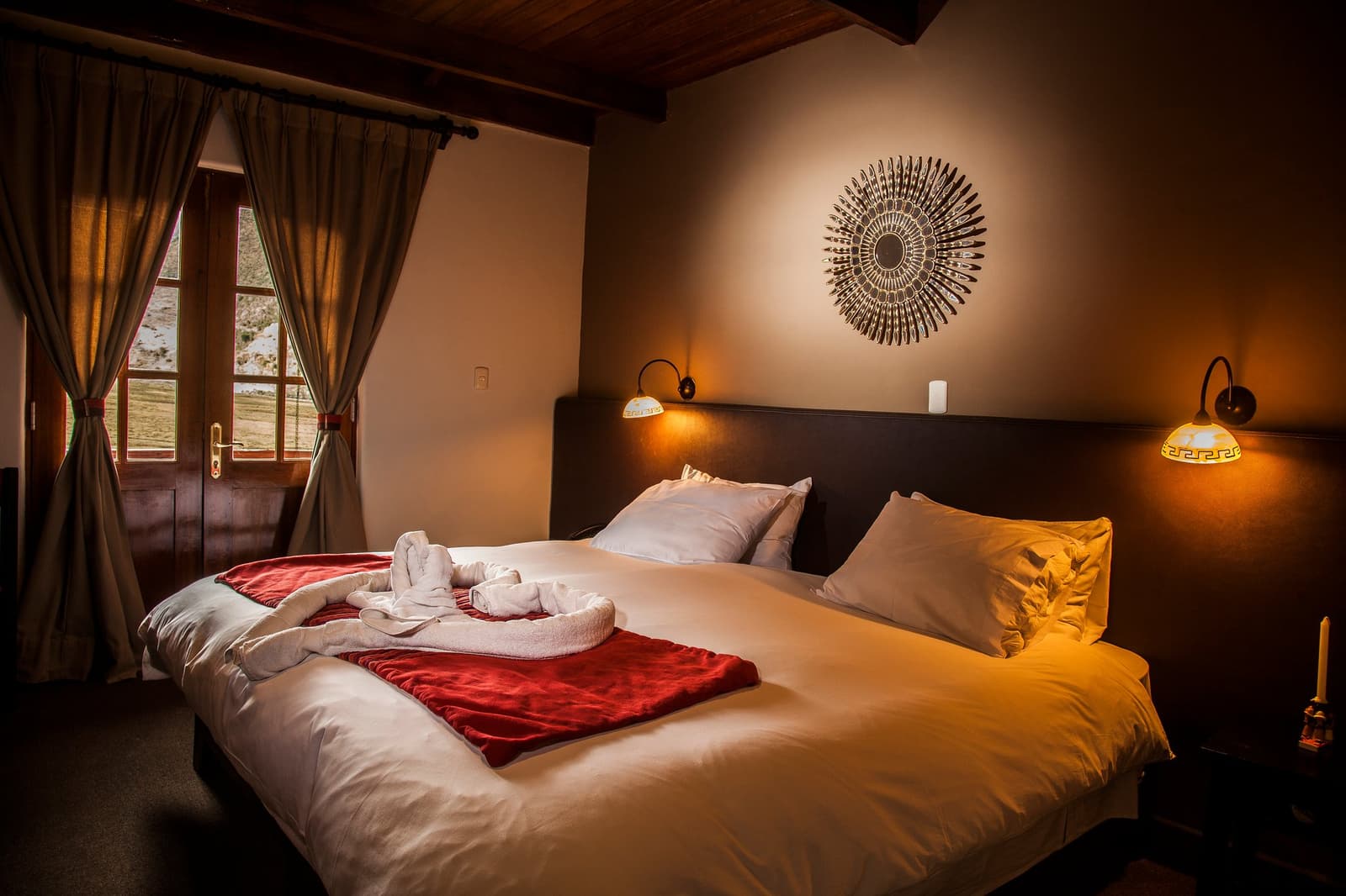 Real beds, warm light, and a valley outside the window. This is the other side of trekking. Warmly lit double bedroom in an Andes mountains tour lodge, featuring a dark wood headboard, white bedding with a red throw, towel art on the bed, wall sconces, a decorative sunburst mirror, and a window overlooking a mountain valley