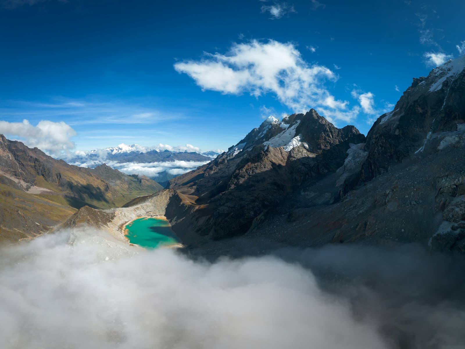 Up here, the clouds are below you and a turquoise lake sits quietly in between. A vivid turquoise glacial lake sits in a high Andean valley on an andes mountains tour, framed by rugged snow-capped peaks and partly veiled by low clouds in the foreground