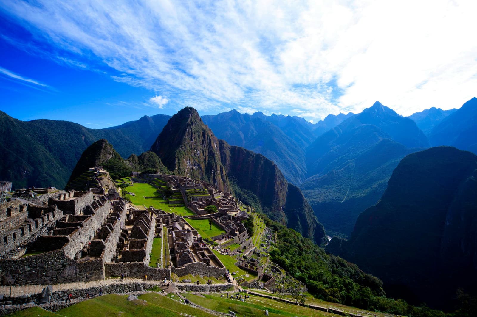 Six hundred years of Inca engineering, still holding its ground. Aerial view of the Machu Picchu citadel on an Andes mountains tour, with Inca stone terraces and ruins spread across a ridge and Huayna Picchu peak rising sharply behind, surrounded by deep forested valleys under a blue sky