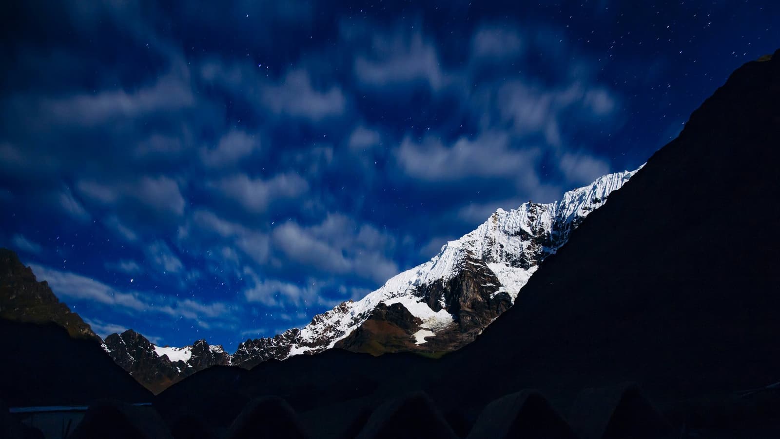 Stars, clouds, and glaciers sharing the same cold sky. Sleep well. Snow-capped Andean peaks glowing under a deep blue night sky scattered with stars and drifting clouds, framed by dark silhouetted ridgelines on this andes mountains tour route in Peru