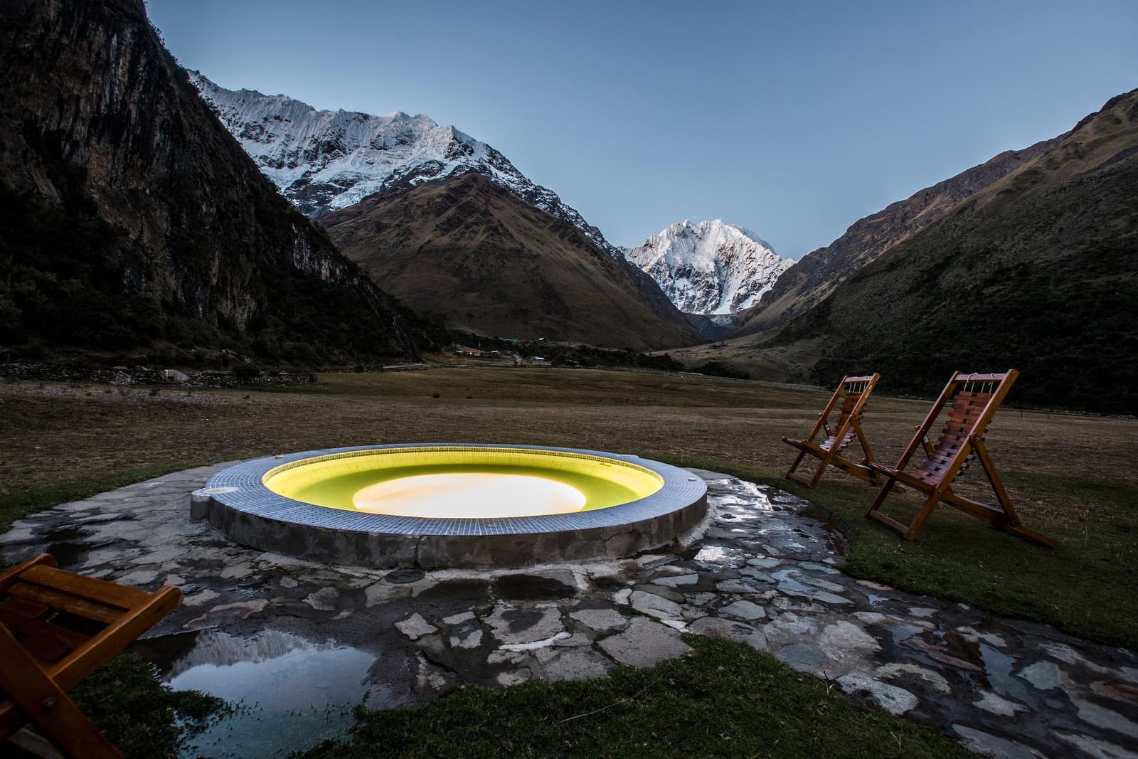 A warm soak at altitude, with the whole valley to yourself. Photo: Alejandro Muller A glowing circular outdoor hot tub on a stone terrace with two wooden deck chairs beside it, set against a high Andean valley framed by snow-capped peaks at dusk, on an Andes mountains tour