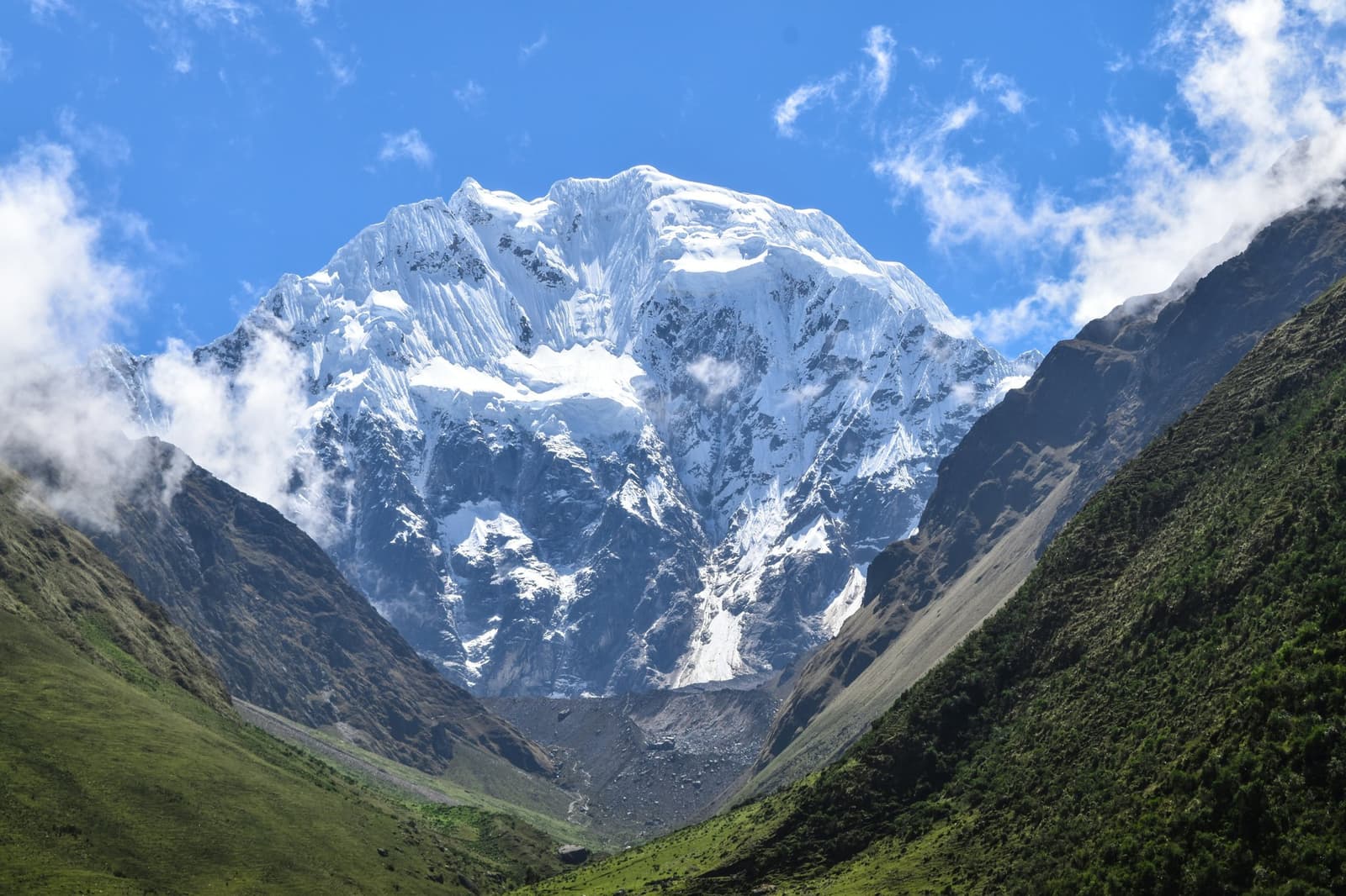 That peak has no interest in your schedule. It was here first, and it shows. A massive snow-capped mountain peak rises above a green glacial valley on an Andes mountains tour in Peru, with rocky scree at its base and wispy clouds around the summit under a vivid blue sky