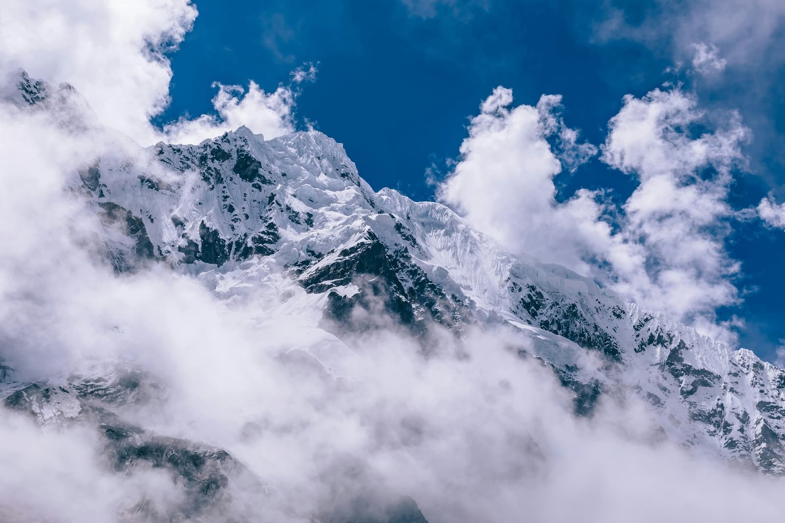 The clouds have opinions about when you get to see the summit. Snow-covered rocky peak emerging through thick white clouds on an andes mountains tour, set against a deep blue sky