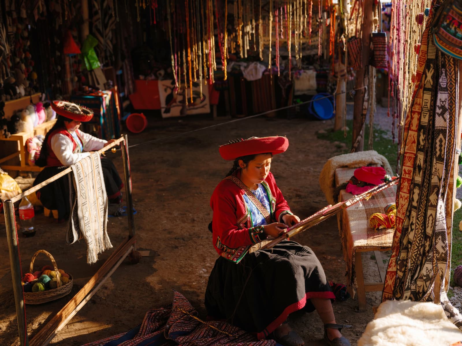 Every thread in that fabric has someone's full attention behind it. Photo: Oleh_Slobodeniuk A woman in traditional red embroidered dress and red hat works a backstrap loom at a colorful Andean textile market, with another weaver and hanging yarn strands visible in the background during an Andes mountains tour