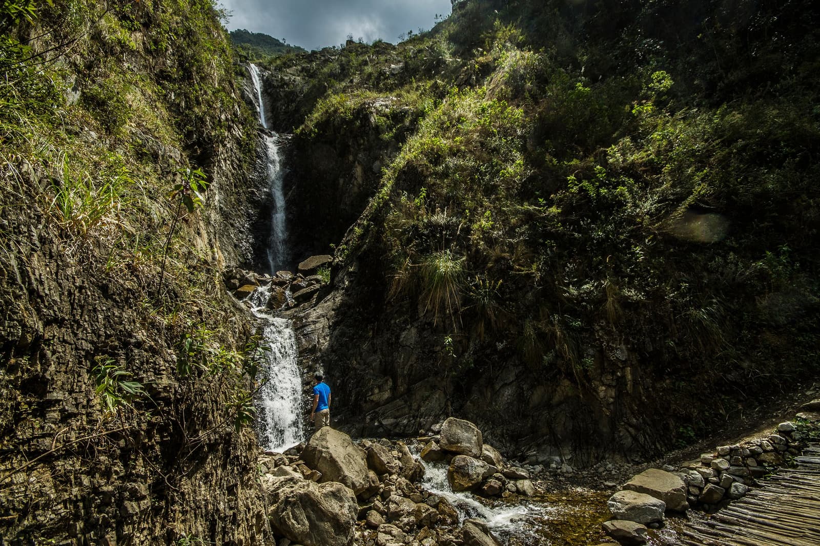 Some stops on the trail have nothing to do with the destination, and everything to do with the moment. Photo: Gustavo Vivanco Leon A lone hiker in a blue shirt stands at the base of a tall two-tiered waterfall cascading through a narrow rocky canyon on an Andes mountains tour, with lush green vegetation covering the steep cliffs and a rustic wooden bridge visible in the foreground
