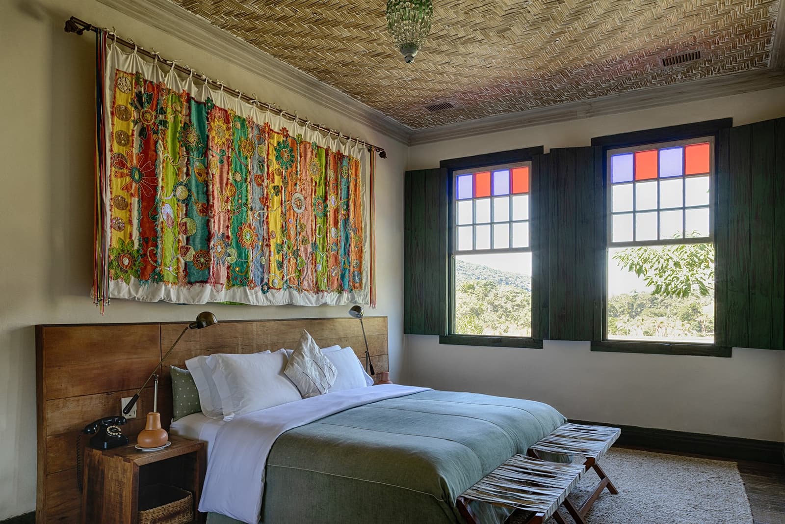 Des murs de ferme centenaires, du linge frais, et la forêt juste là. Photo: Henrique Mangeon Une chambre d'hôte du lodge Engenho de l'Ibiti Project, avec un lit double à tête de lit en bois habillé de linge blanc et d'un jeté sauge, un grand textile brodé et coloré accroché au mur, un plafond tressé et deux fenêtres de style colonial avec des vitraux rouges et bleus donnant sur des collines boisées