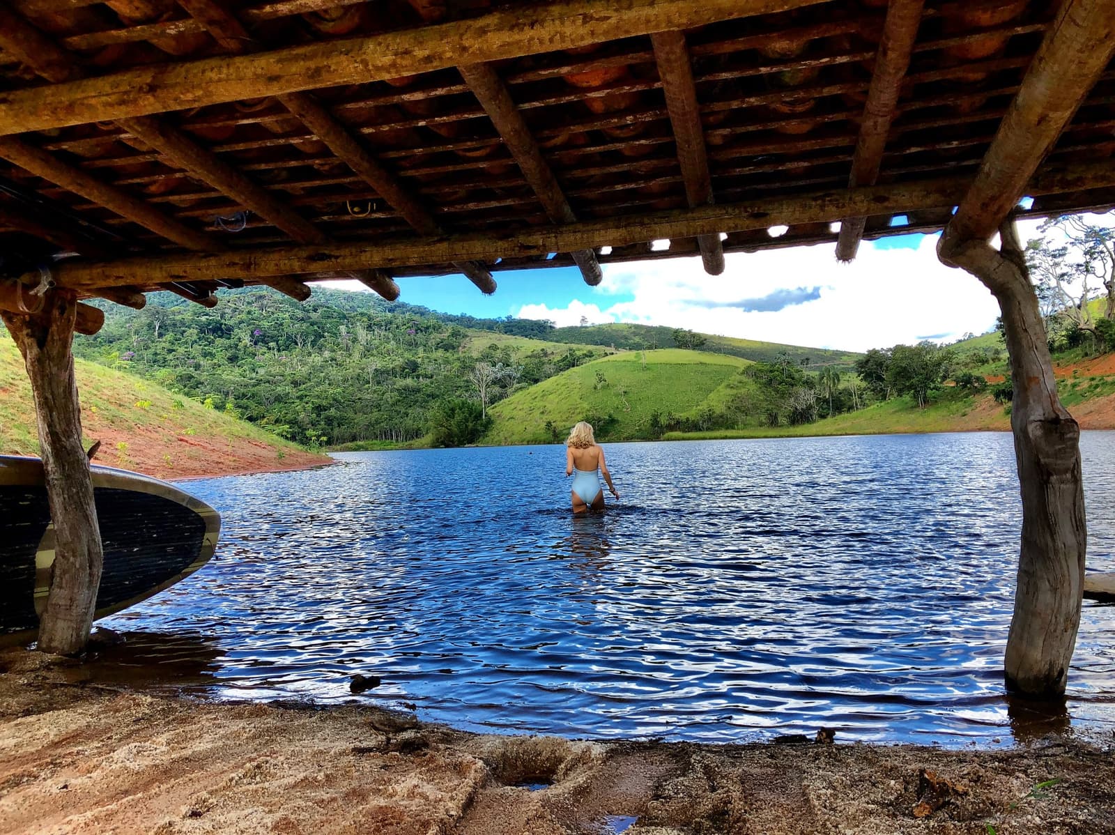 Lago Negro an einem sonnigen Nachmittag. Ein paar Schritte weiter lohnen sich. Eine Frau im hellblauen Badeanzug watet in einen ruhigen See im Ibiti Project, gerahmt von rustikalen Holzpfeilern und einem Schuppendach, im Hintergrund grüne Hügel und Atlantischer Regenwald