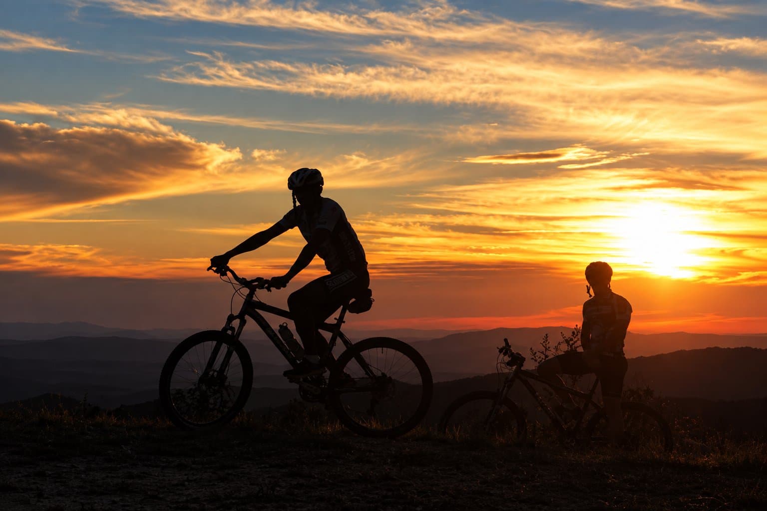 Les jambes font le trajet, le ciel fait le spectacle. Deux cyclistes en silhouette sur fond de coucher de soleil orange et rouge intense au-dessus de crêtes montagneuses, lors d'une sortie à l'Ibiti Project