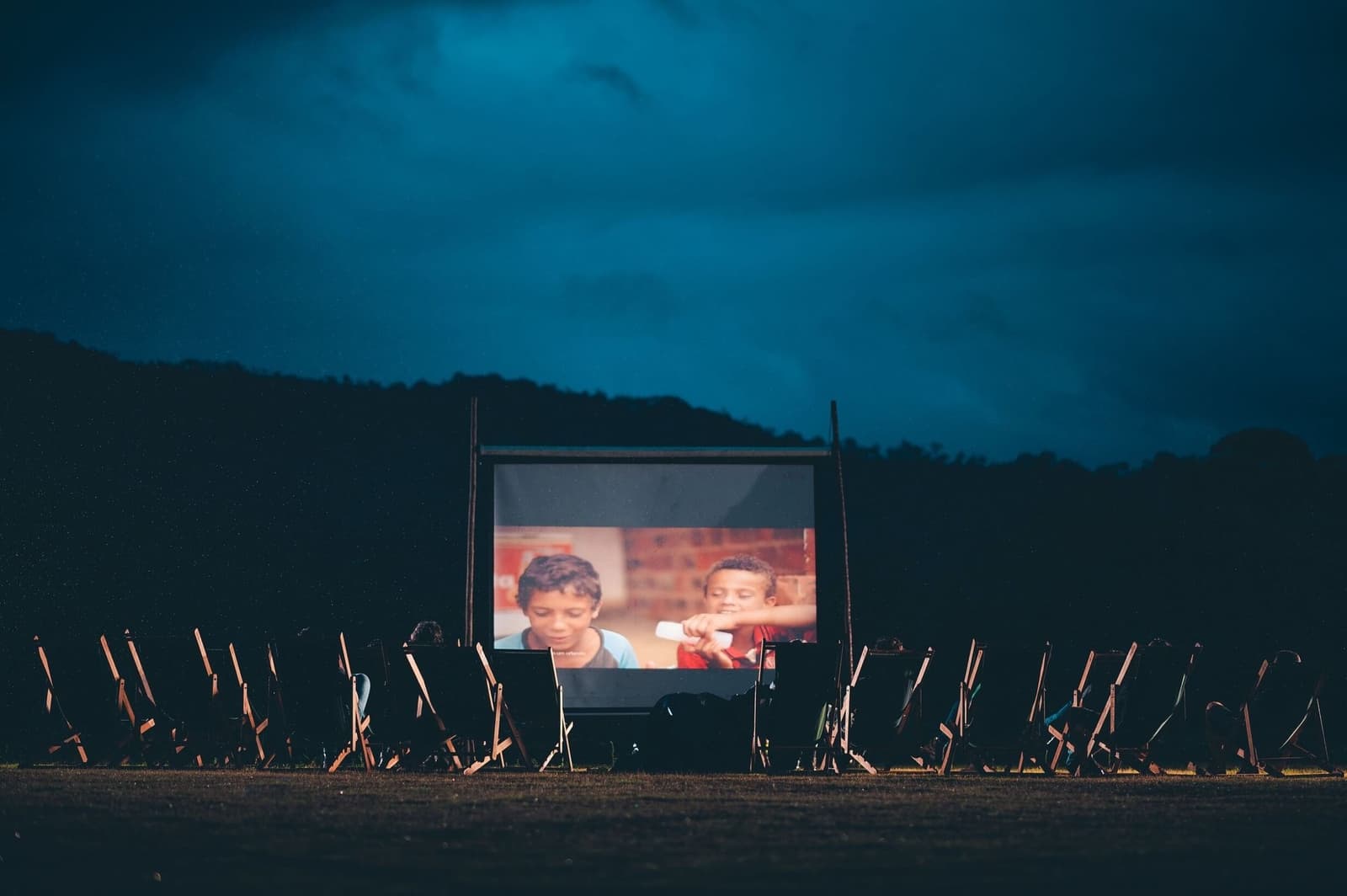 Cinéma en plein air, forêt derrière vous, rien d'autre à prévoir. Des rangées de chaises pliantes en bois vides font face à un écran de cinéma en plein air illuminé dans l'obscurité, projetant deux enfants dans une scène de film, avec une colline boisée et un ciel bleu nuageux en arrière-plan, dans le cadre de l'Ibiti Project.