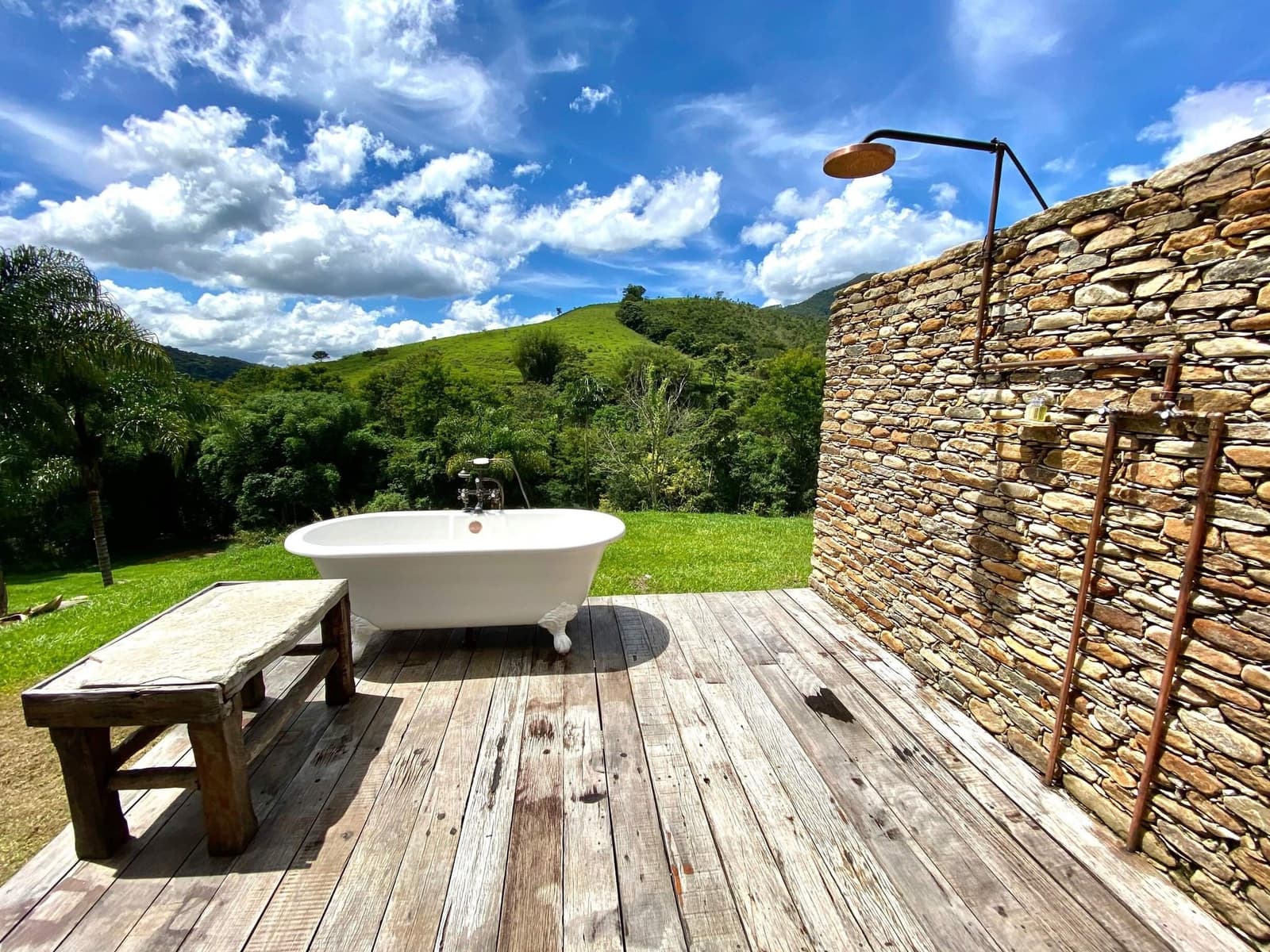 Un bain en plein air, les collines en guise de rideau. Une baignoire sur pieds blanche posée sur une terrasse en bois brut à l'Ibiti Project, avec une douche en cuivre contre un mur de pierre sèche et des collines verdoyantes sous un ciel bleu