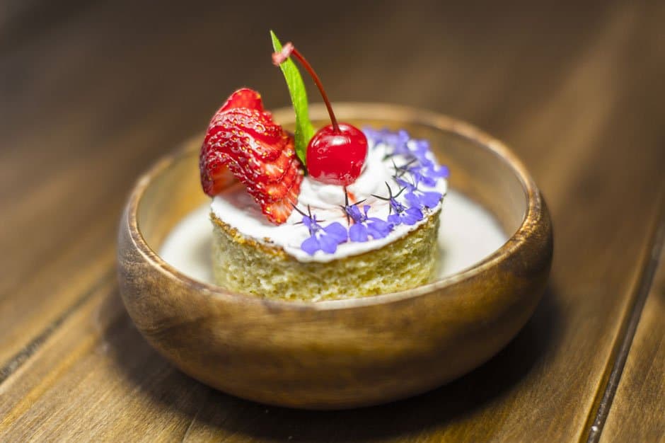 Proof that a wooden bowl can hold something genuinely elegant. A small sponge cake topped with whipped cream, a glazed cherry, sliced strawberry, and delicate purple edible flowers, served in a rustic wooden bowl on a wood-plank table — the kind of plated dessert you find on a luxury Machu Picchu tour that takes local ingredients seriously.