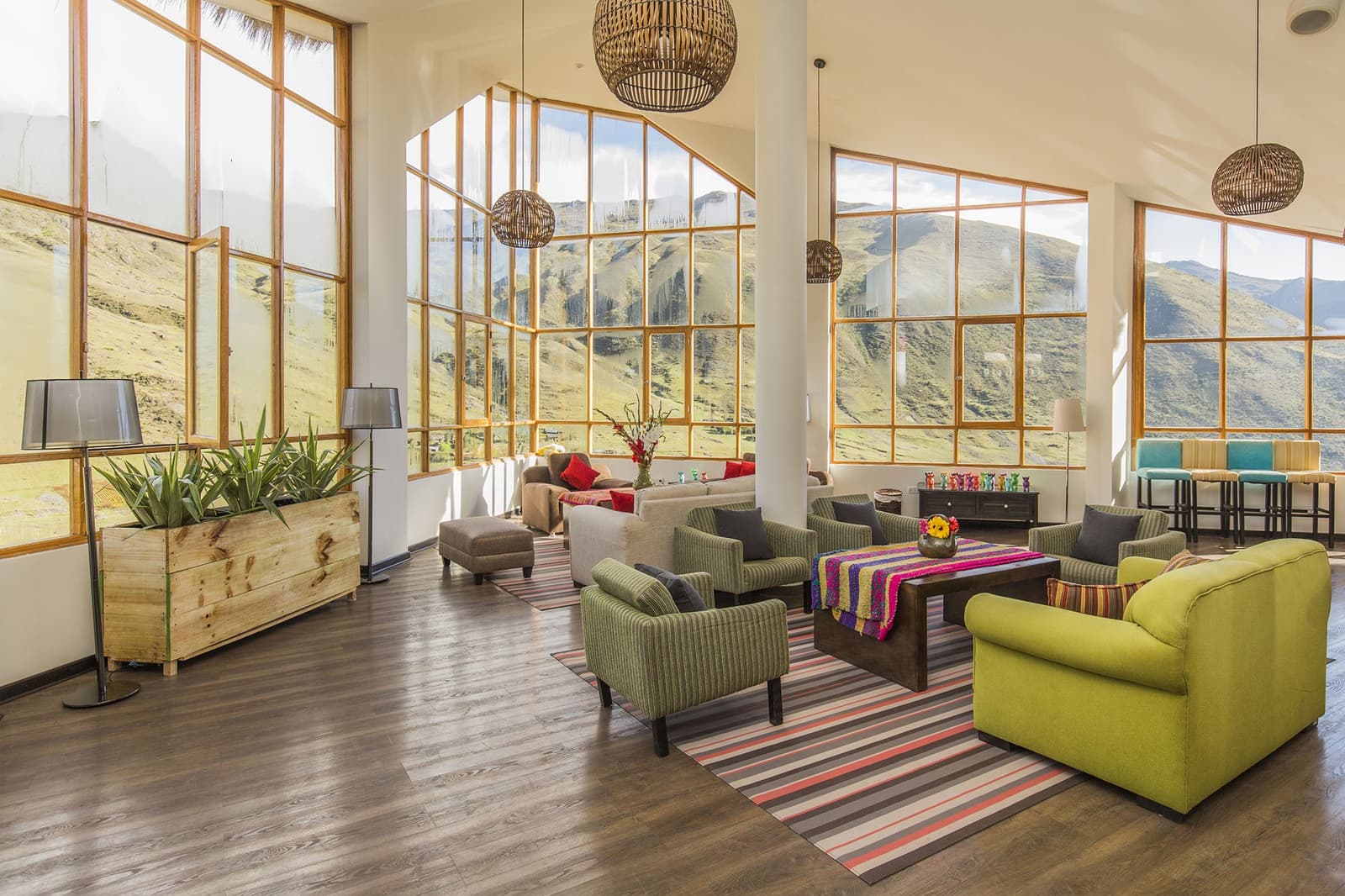 The mountains are right there, and the armchairs are surprisingly comfortable. Bright open lounge of a high-altitude Andean lodge on a luxury Machu Picchu tour, featuring colorful upholstered armchairs, a striped Peruvian textile table runner, wicker pendant lights, and floor-to-ceiling wooden-framed windows overlooking golden-green mountain slopes