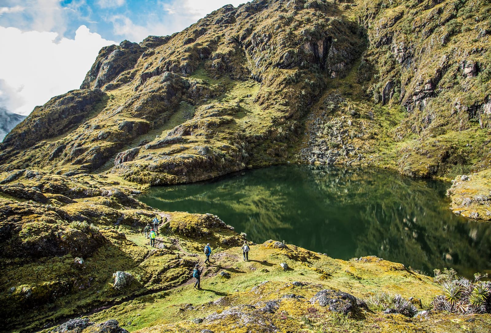 Four hours of trail, one lake that earns its silence. A small group of hikers follows a narrow trail around a deep green glacial lake on this luxury Machu Picchu tour, surrounded by moss-covered rocks and high Andean peaks under a partly cloudy sky