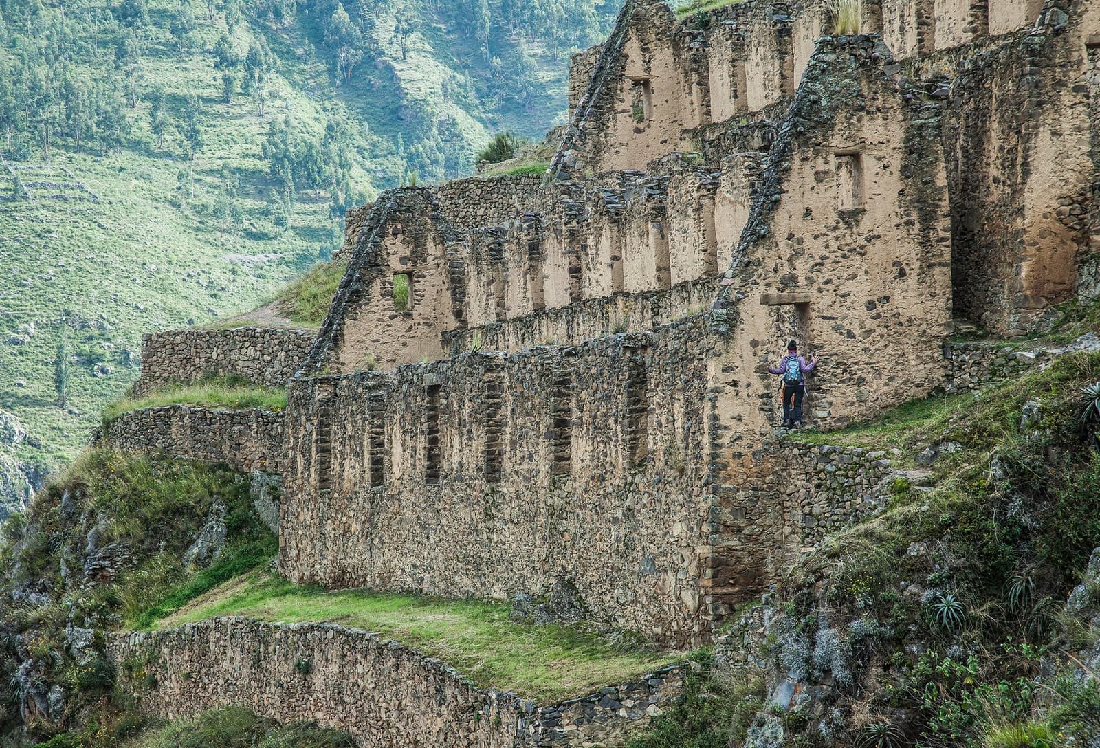 Rarely visited, quietly enormous. The storehouses kept an empire fed. A lone hiker with a backpack stands beside the towering multi-tiered stone walls of ancient Inca storehouses on a steep Andean hillside, part of a luxury Machu Picchu tour itinerary, with terraced ruins descending toward a green mountain valley below