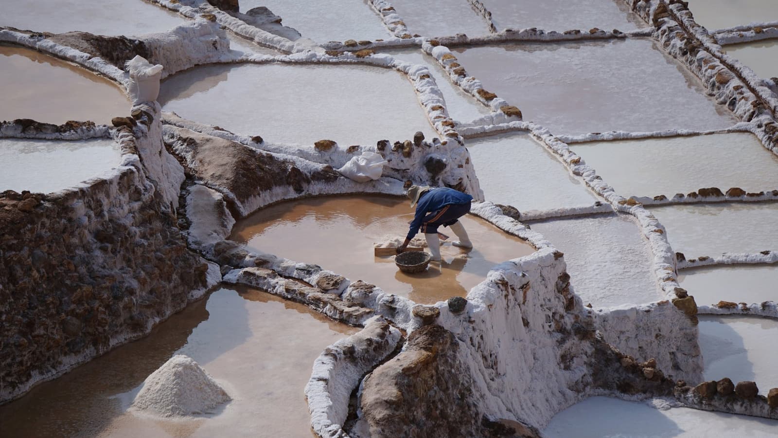 The same pools, the same method, working since Inca times. Some things just hold. A worker in a blue jacket bends to harvest salt by hand at the Maras salt pans, surrounded by dozens of white mineral-crusted terraced pools on this luxury Machu Picchu tour itinerary