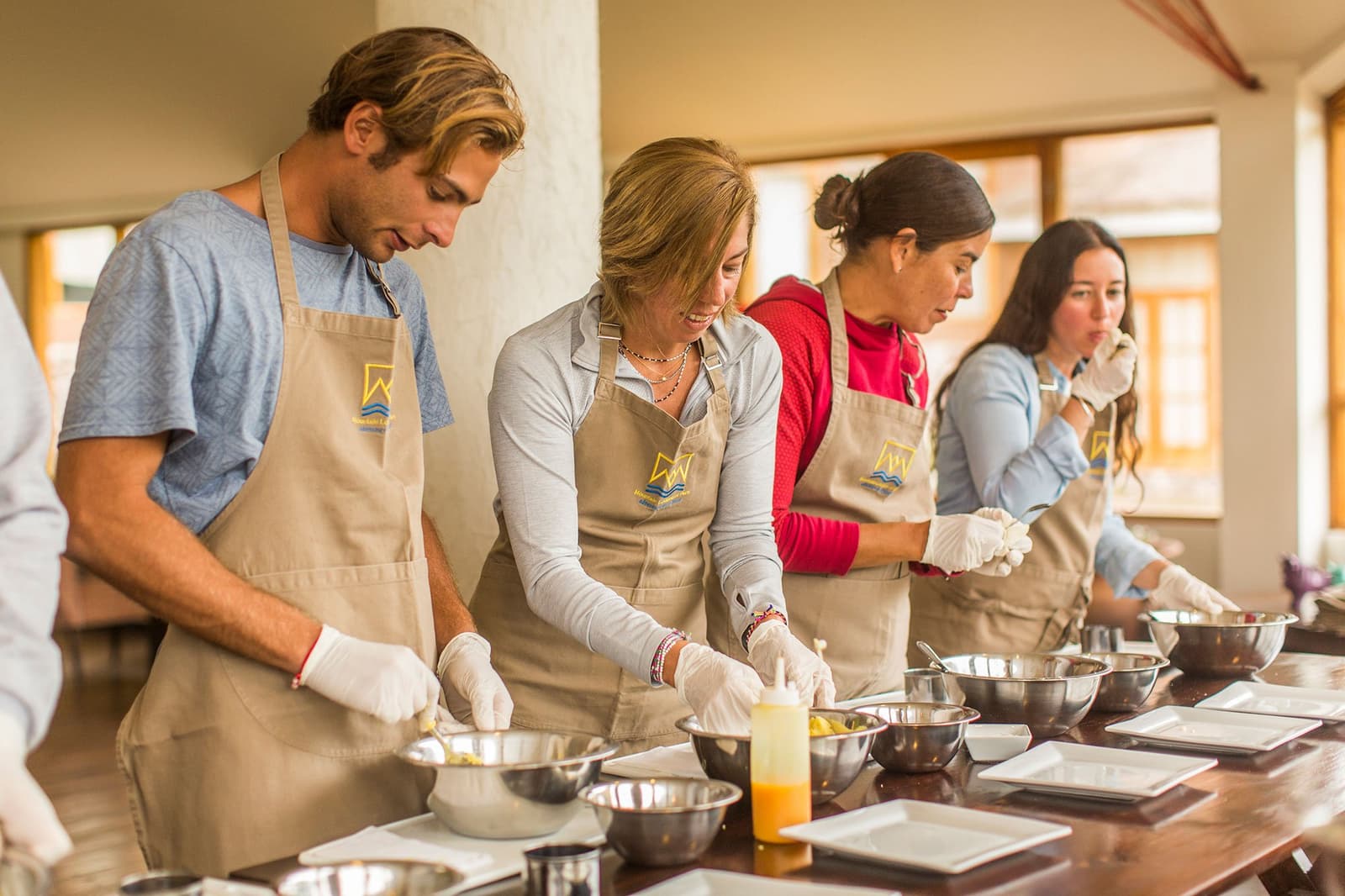 Hands in the bowl, gloves on, everyone suddenly an expert. The Andes make good students of us all. Four travelers wearing branded aprons and food-safe gloves prepare dishes with mixing bowls and ingredients at a long wooden table during a Peruvian cooking class on a luxury Machu Picchu tour