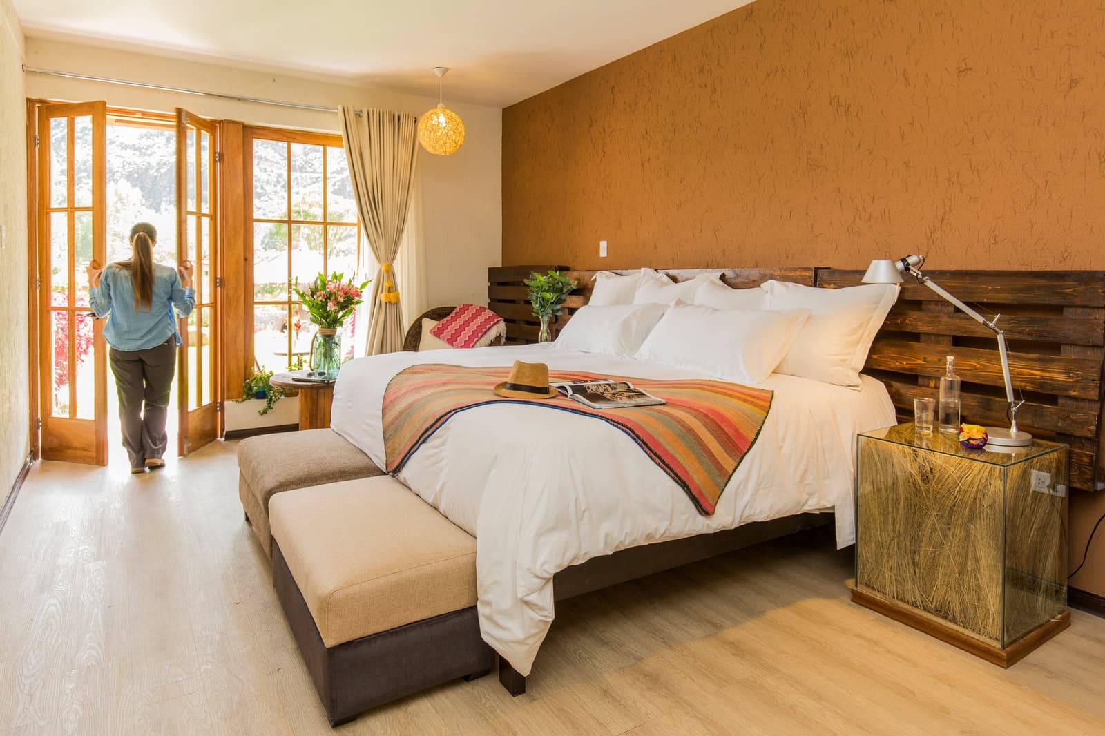 A hat on the pillow, flowers on the table, and the Andes right outside the door. Bright lodge bedroom on a luxury Machu Picchu tour featuring a king bed with a striped Andean throw, reclaimed-wood headboard, woven pendant light, and a guest opening folding wooden doors to a garden view
