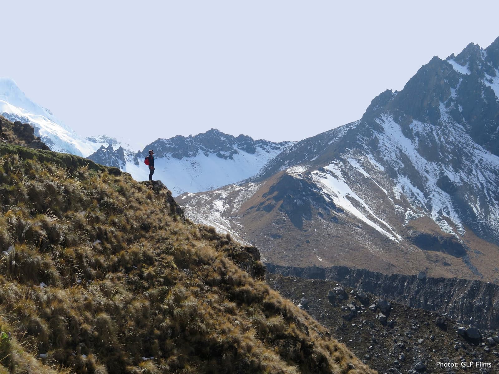 Small figure, enormous mountain. That's the whole deal up here. Photo: GLP Films A lone hiker with a red backpack stands on a rocky ridge covered in tussock grass on the luxury Salkantay trek, with snow-streaked Andean peaks filling the horizon behind them