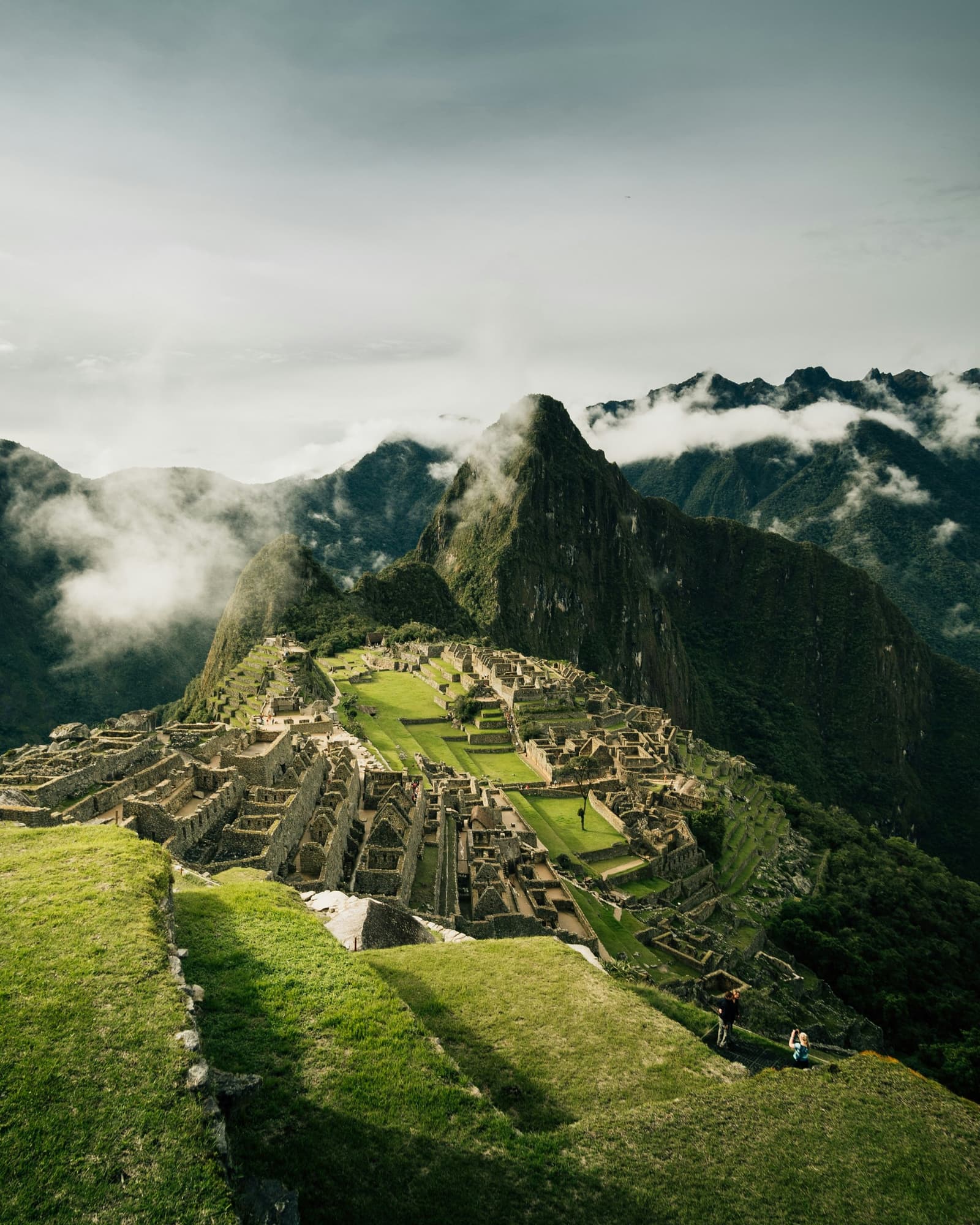 Stone walls, cloud-wrapped peaks, and the quiet realisation that you walked here. Aerial view of the Inca citadel of Machu Picchu on a cloudy morning, with Huayna Picchu peak rising behind the stone ruins and two visitors sitting on a grassy terrace in the foreground, seen at the end of the luxury Salkantay trek
