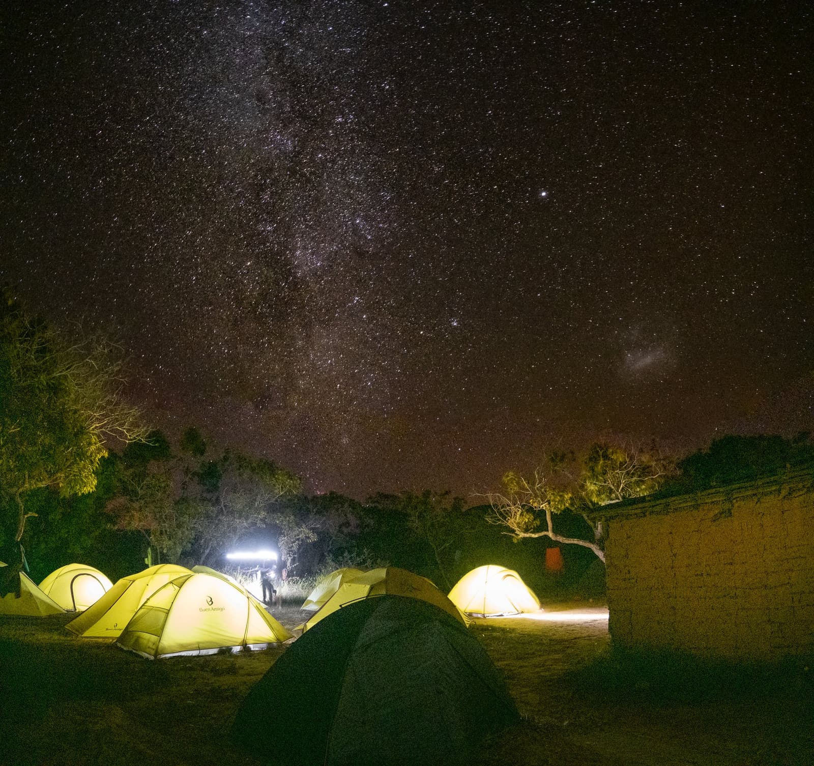 Longe o suficiente da cidade para a Via Láctea aparecer de verdade. Tendas amarelas iluminadas montadas num acampamento durante uma expedição ao Monte Roraima, com a Via Láctea atravessando um céu estrelado sobre árvores esparsas e uma parede de adobe