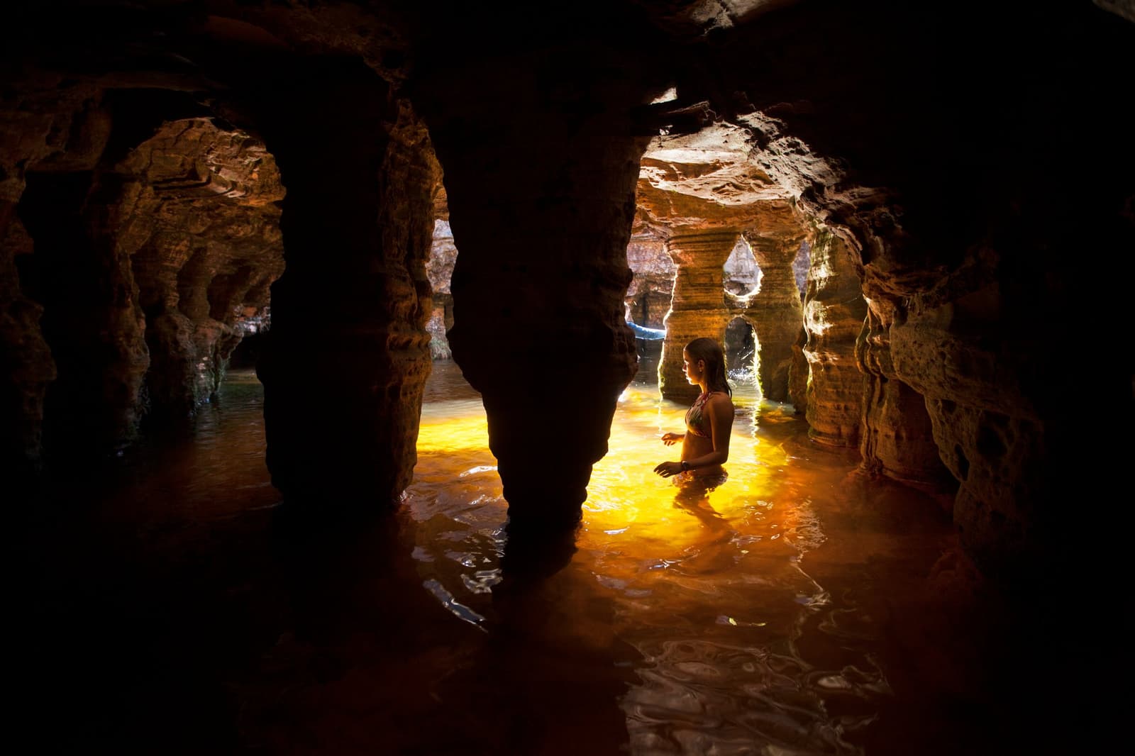Água fresca, luz quente, e uma caverna que levou dois bilhões de anos para ser esculpida. Uma mulher caminhando até a cintura em uma caverna de arenito inundada durante uma expedição ao Monte Roraima, com luz dourada refletida na água cor de âmbar entre colunas de rocha esculpidas