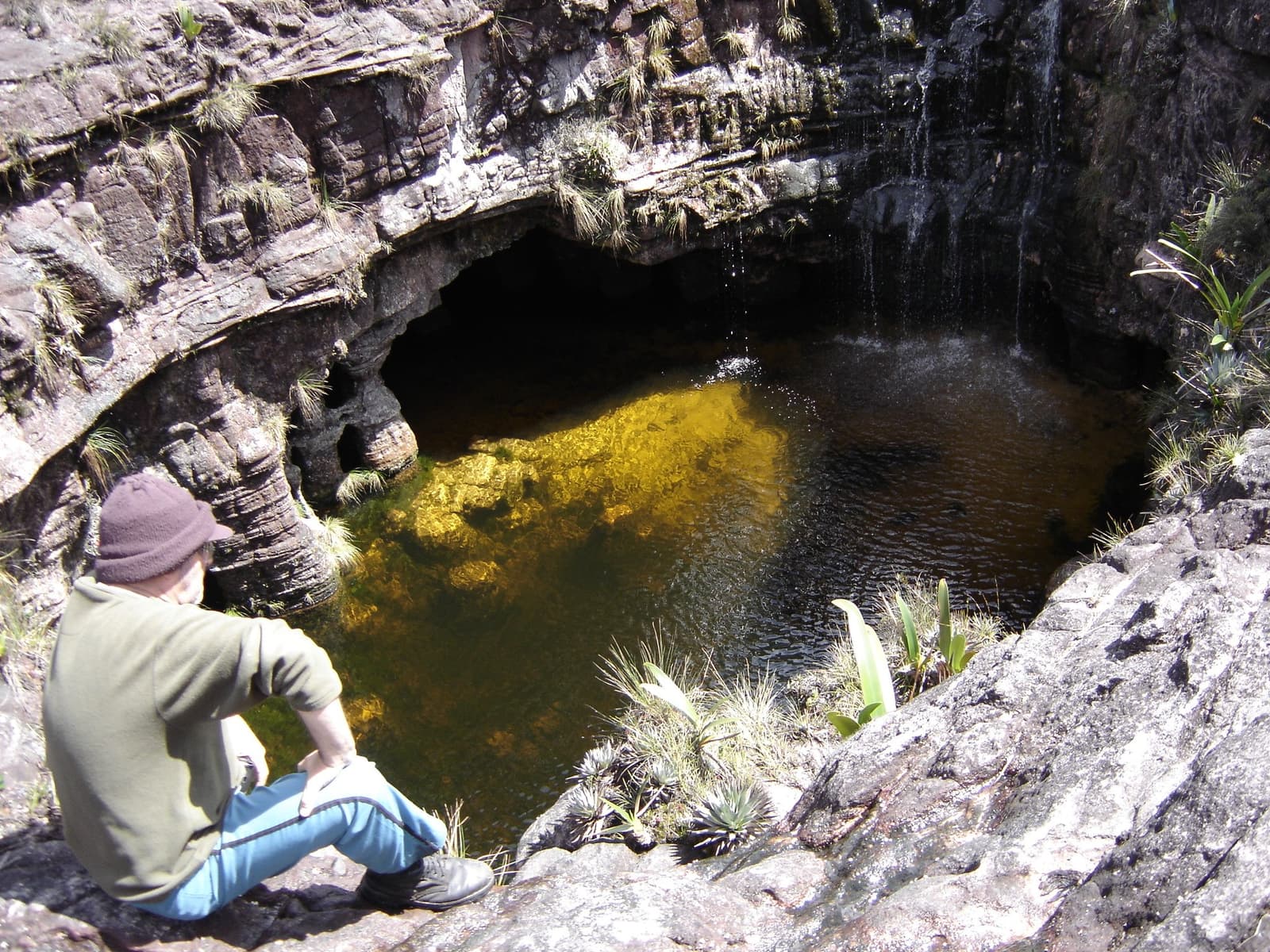 Uma piscina assim faz qualquer um esquecer os quilômetros que ainda faltam. Um caminhante de gorro e moleton sentado à beira de uma piscina natural circular em rocha, alimentada por uma fina queda d'água, com a água cor de âmbar brilhando dourada onde a luz do sol ilumina o fundo coberto de algas, cercado por paredes de tepui e bromélias silvestres durante uma expedição ao Monte Roraima