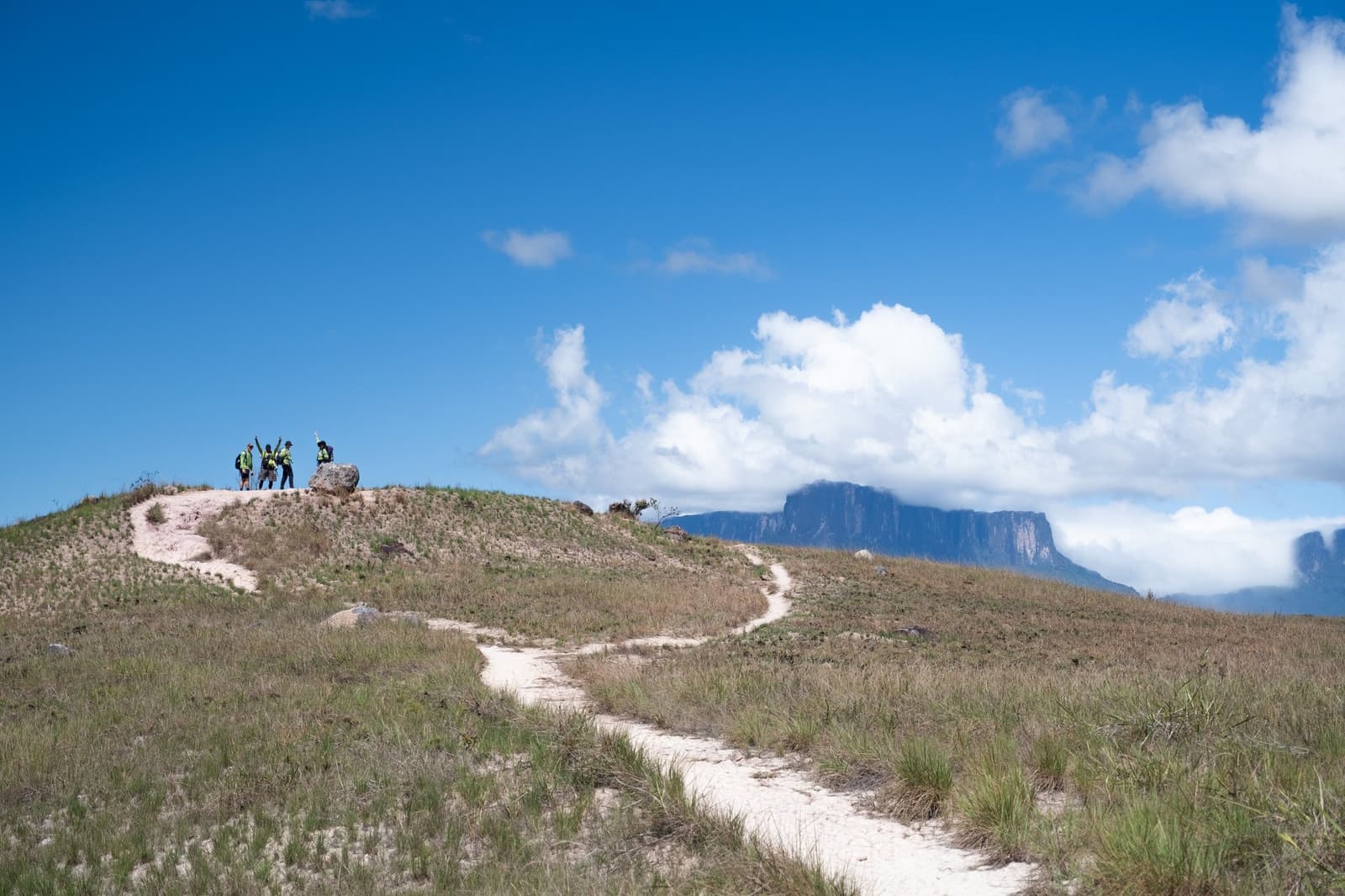 Sempre tem alguém que levanta os braços primeiro, e aí os outros não resistem. Quatro caminhantes comemoram no topo de uma colina arenosa durante a expedição ao Monte Roraima, com o tepui de topo plano visível entre as nuvens ao fundo