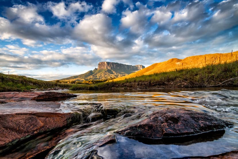 A shallow rocky river flows toward a flat-topped tepui on a mount roraima expedition, with golden savanna hills and a wide cloudy sky above A shallow rocky river flows toward a flat-topped tepui on a mount roraima expedition, with golden savanna hills and a wide cloudy sky above
