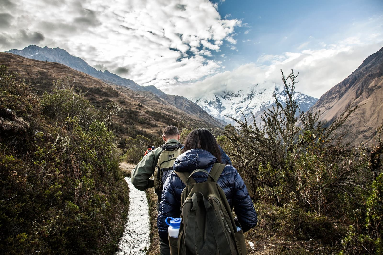 The path does the persuading, one step at a time. Two hikers with backpacks walk a narrow trail on a salkantay tour, with scrubby Andean vegetation on both sides and snow-capped peaks rising under a dramatic cloudy sky ahead