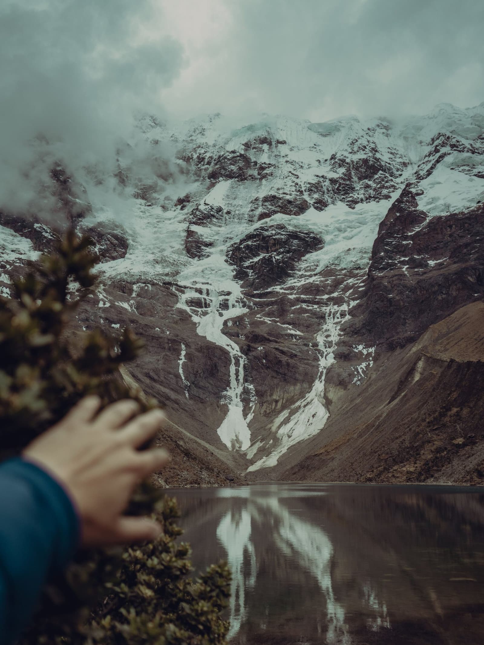 Cold water, colder peaks, and the glacier doing all the work. A traveler's hand reaches toward alpine scrub in the foreground while a snow-streaked glacier feeds into a calm glacial lake below towering Andean peaks on an Andes mountains tour in Peru