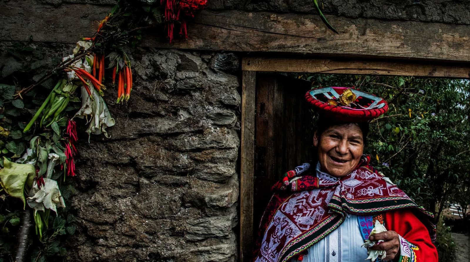 She opened the door, and honestly, that was the warmest welcome of the whole trip. A smiling Quechua woman in traditional red woven cloak and embroidered hat stands in a stone doorway during an Andes mountains tour, with wild flowers hanging on the wall beside her