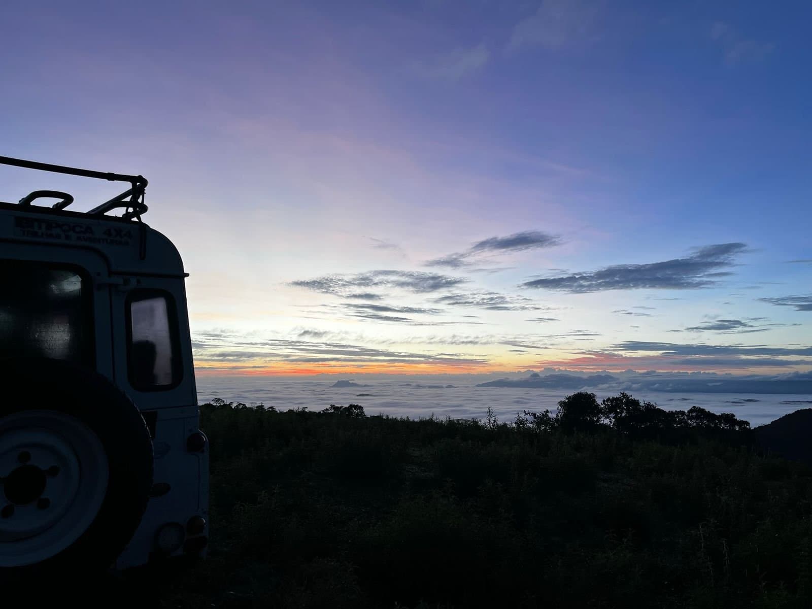 Früher als das Tal wach, und über den Wolken geparkt. Silhouette eines 4x4-Geländefahrzeugs in der Morgendämmerung über einem Wolkenmeer, mit einem warm gefärbten Horizont über dem Hochland des Ibiti Project